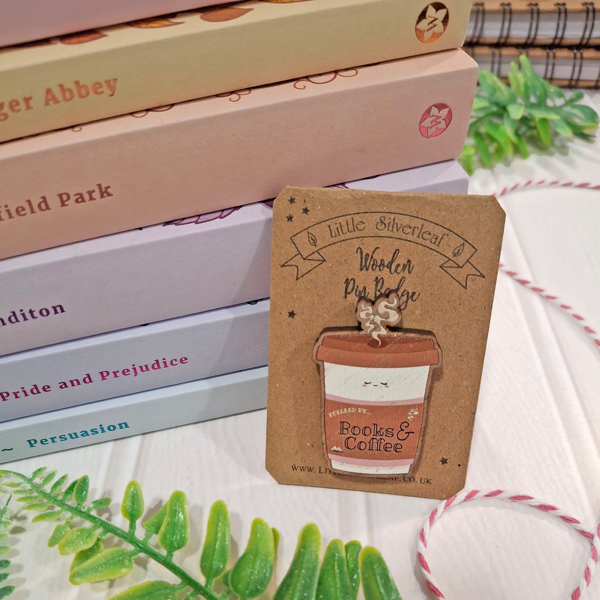 A small wooden books-and-coffee pin badge displayed on its kraft backing card. The card is styled on a white tabletop next to stacked books, green leaves, and pink-and-white string.