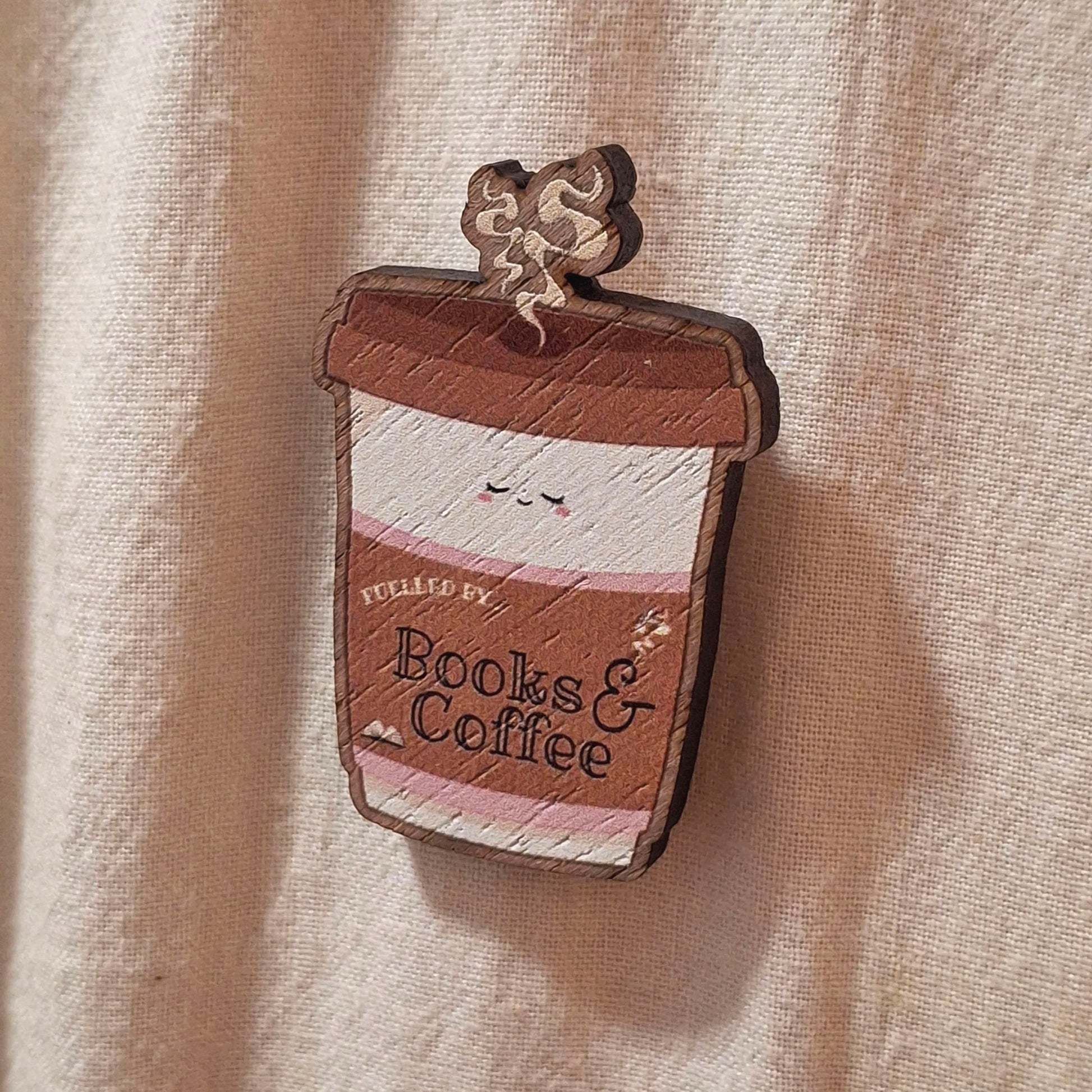 A small wooden pin badge shaped like a takeaway coffee cup with the words 'Fuelled by Books & Coffee' on the front, photographed against a cream fabric tote bag background.