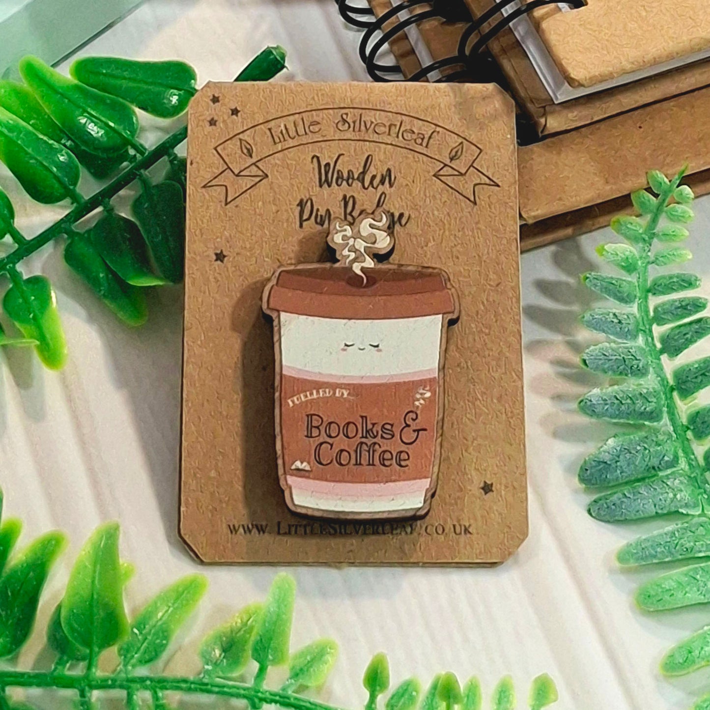 A wooden pin badge shaped like a coffee cup with the text 'FUELLED BY Books & Coffee', displayed on a kraft backing card. The badge is styled with green leaves and a notebook in the background.