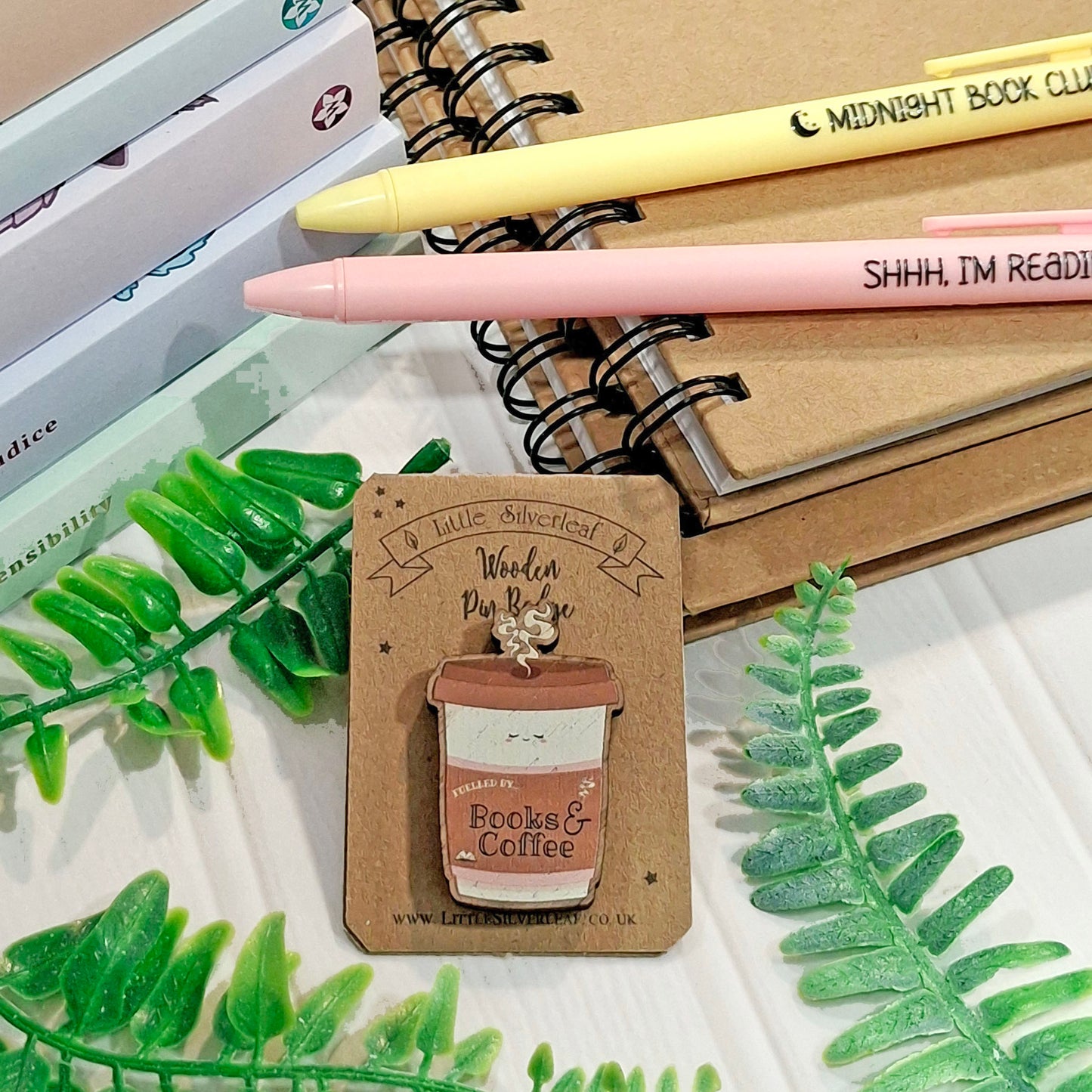 A cute coffee cup pin badge with a 'fuelled by books and coffee' slogan presented on a backing card, styled on top of notebooks and pens with decorative green leaves.