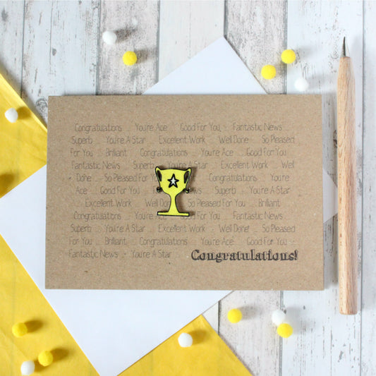 A handmade brown kraft card featuring a yellow 3D trophy embellishment, the word 'Congratulations' printed in black, and other congratulatory phrases printed in the background.
