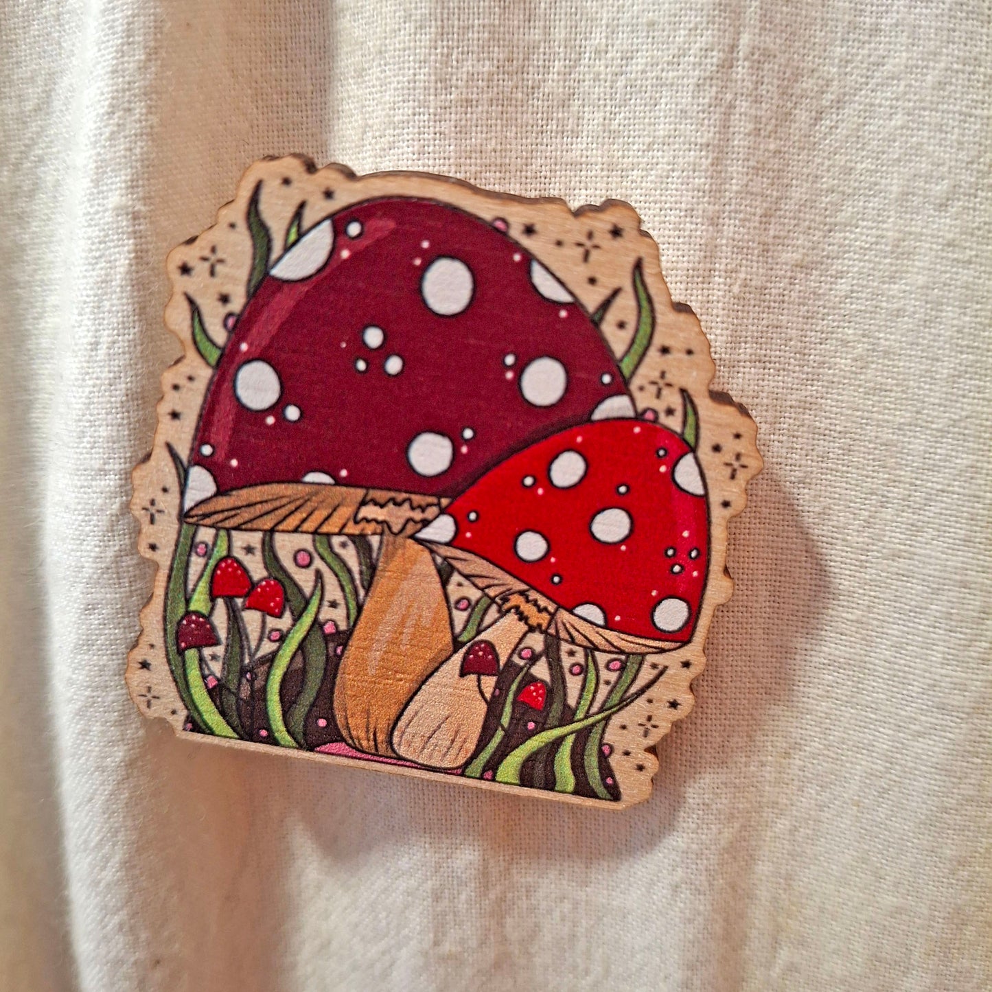 A wooden pin shaped like a cluster of red and burgundy mushrooms with white spots, green stems, and a scalloped border, displayed on a textured beige fabric background.