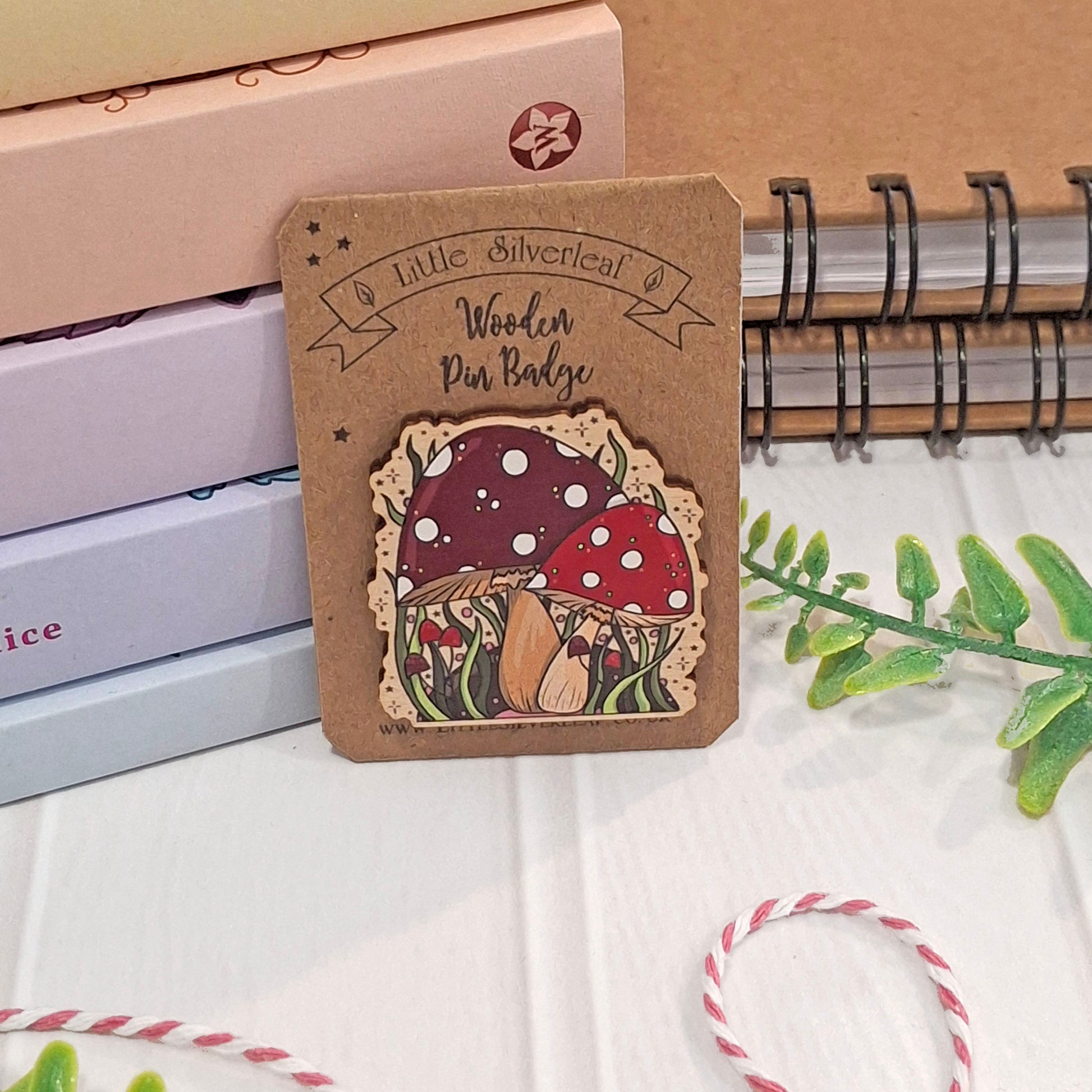 A wooden pin badge featuring a cute mushroom design, mounted on a kraft backing card. The badge is styled on a white surface with a brown notebook, books, and green leaves in the background.