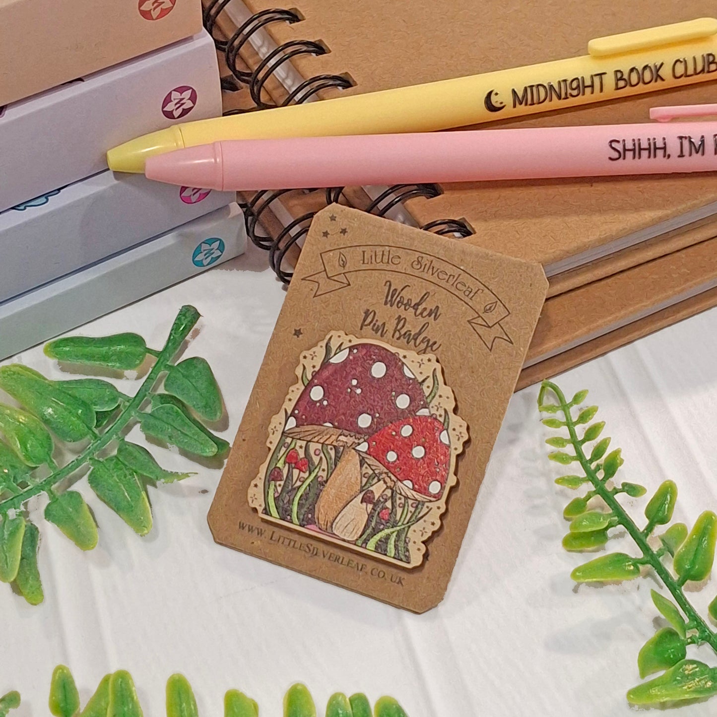 A wooden pin badge featuring red mushrooms with white spots, photographed on a white surface with notebooks, pens, and green leaves around it.