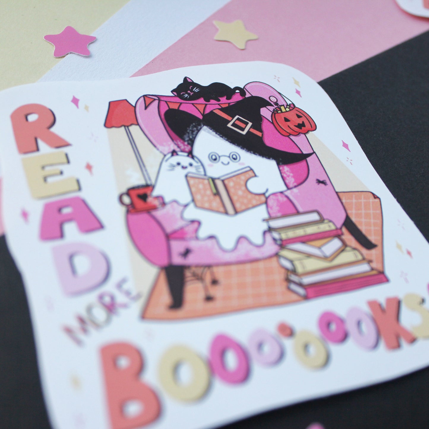 A close-up of a large vinyl sticker featuring two ghosts, the older one wearing a witch hat and reading books to the younger one, with the words “READ MORE BOOOOOOKS” in colorful lettering. The product is photographed on a styled flat-lay background with pastel paper and star shapes.