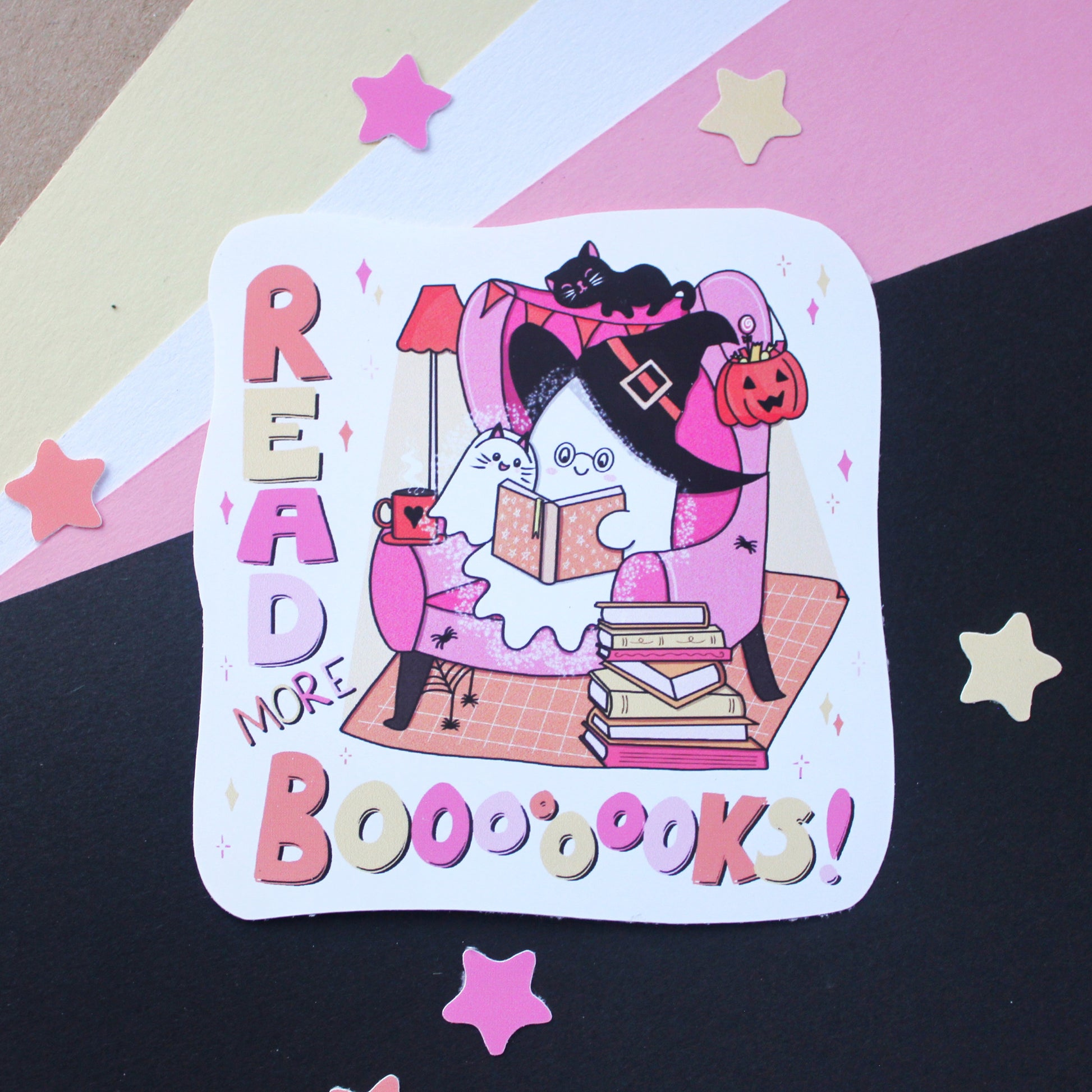 A large vinyl die-cut sticker featuring a whimsical Halloween illustration of two ghosts, reading a book on a pink armchair with a pumpkin, black cat, and books around them. The text reads 'READ MORE BOOOOKS!' in playful pastel lettering.