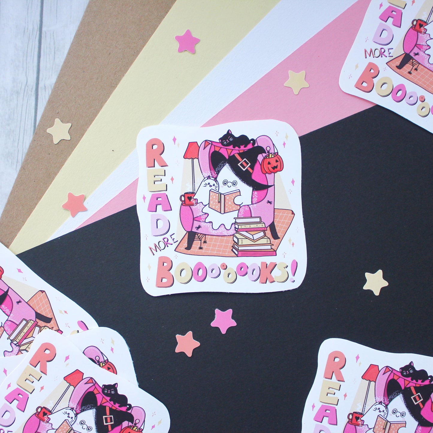 A square sticker with rounded corners featuring a whimsical illustration of a ghost dressed as a witch reading a book to a ghost dressed as a cat, surrounded by the words 'READ MORE BOOBOOKS!' in pink and orange lettering. The sticker is displayed on a styled flat-lay background of black, pink, beige, and white paper with scattered star cutouts.