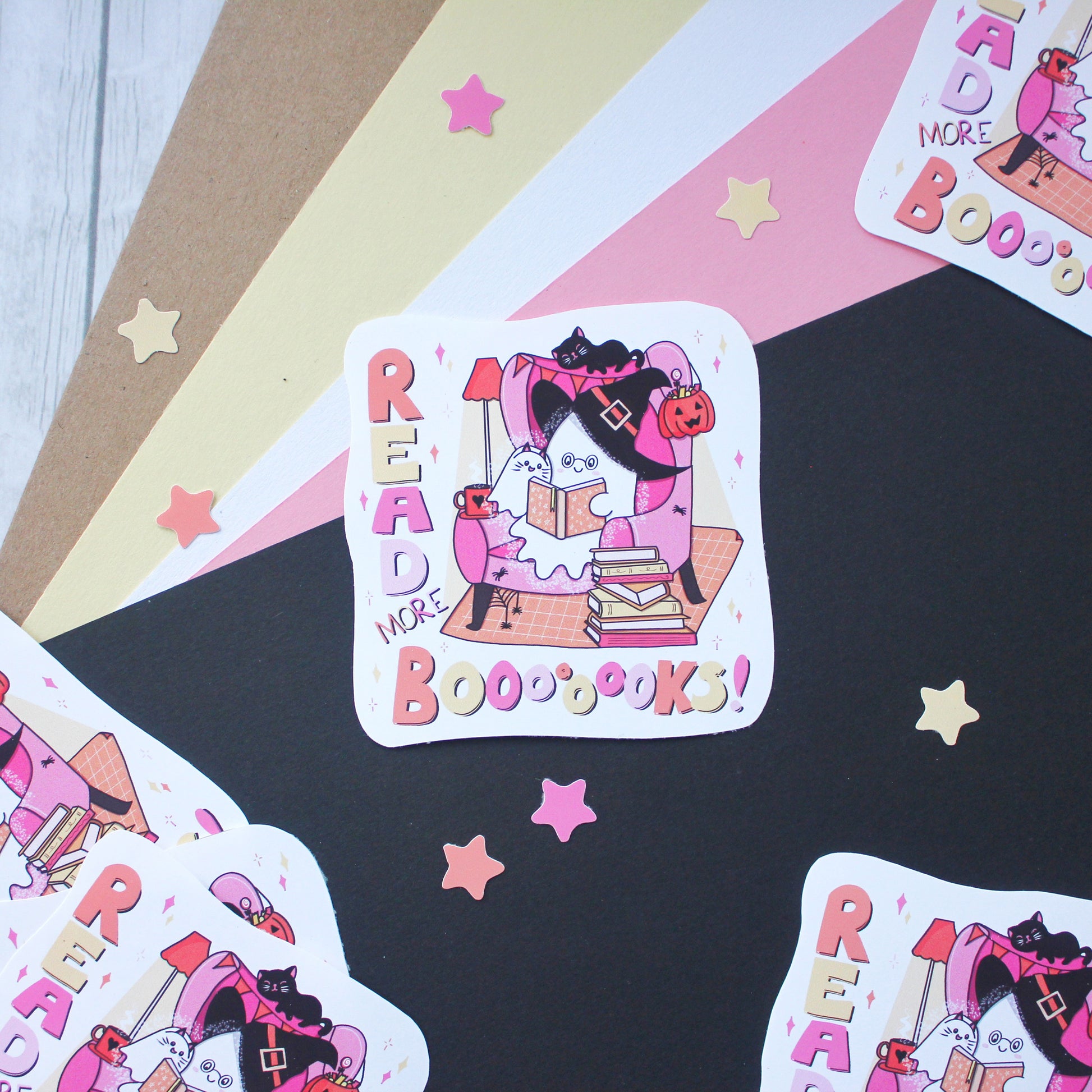 A square sticker with rounded corners featuring a whimsical illustration of a ghost dressed as a witch reading a book to a ghost dressed as a cat, surrounded by the words 'READ MORE BOOBOOKS!' in pink and orange lettering. The sticker is displayed on a styled flat-lay background of black, pink, beige, and white paper with scattered star cutouts.