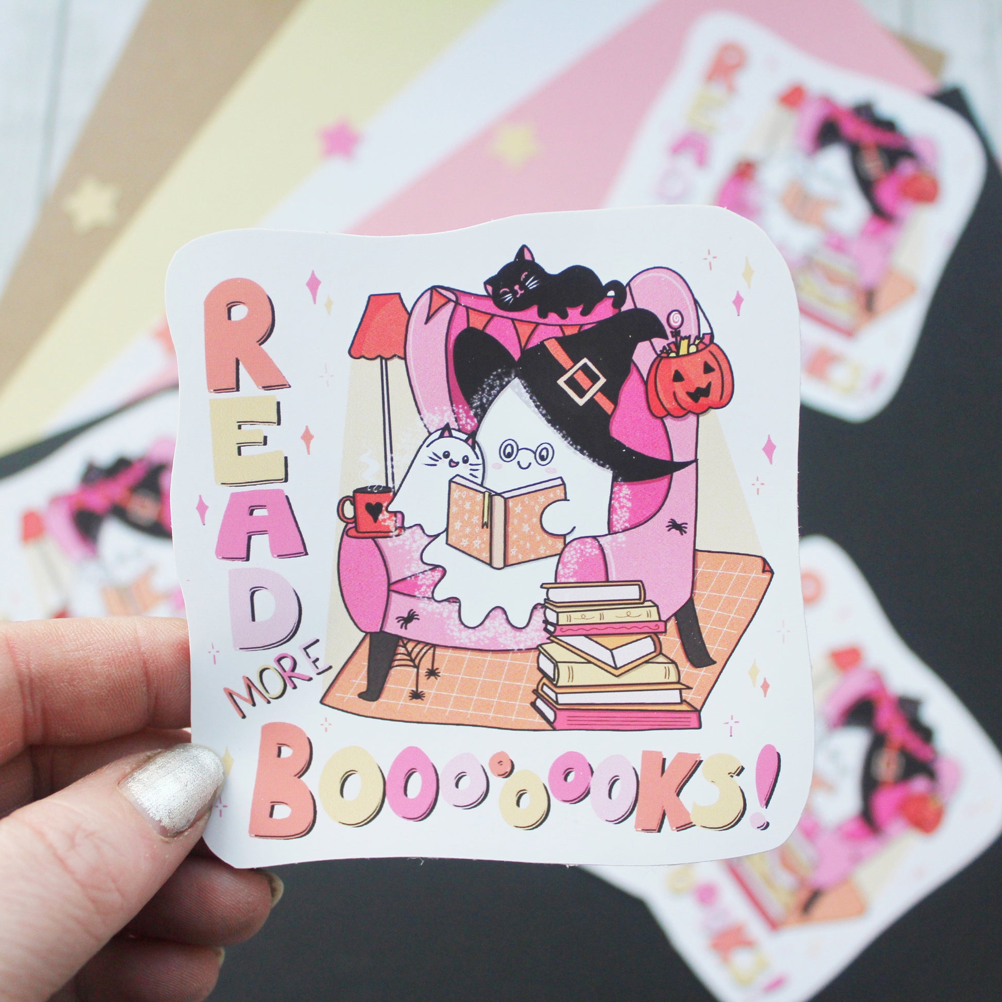 A hand holds a die-cut sticker featuring a whimsical Halloween illustration of ghosts, a cat, and books, with the text 'READ MORE BOOOOKS!'. Other similar stickers are blurred in the background.