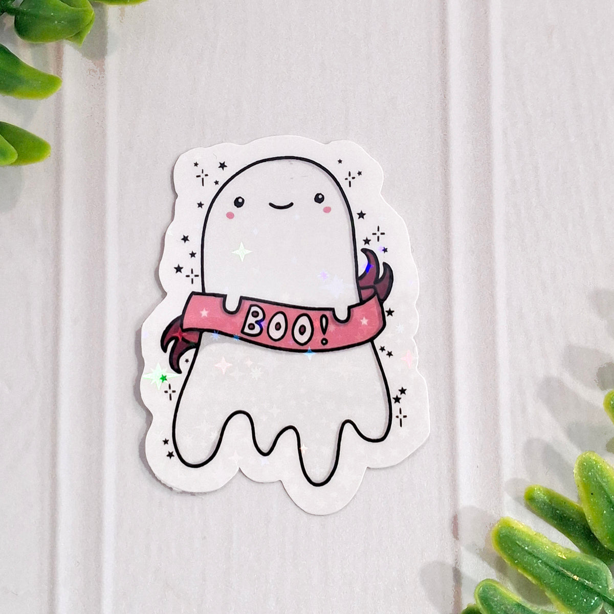 A cute holographic ghost sticker with a pink 'BOO!' banner, placed on a white wood background with green plant leaves in the corners.