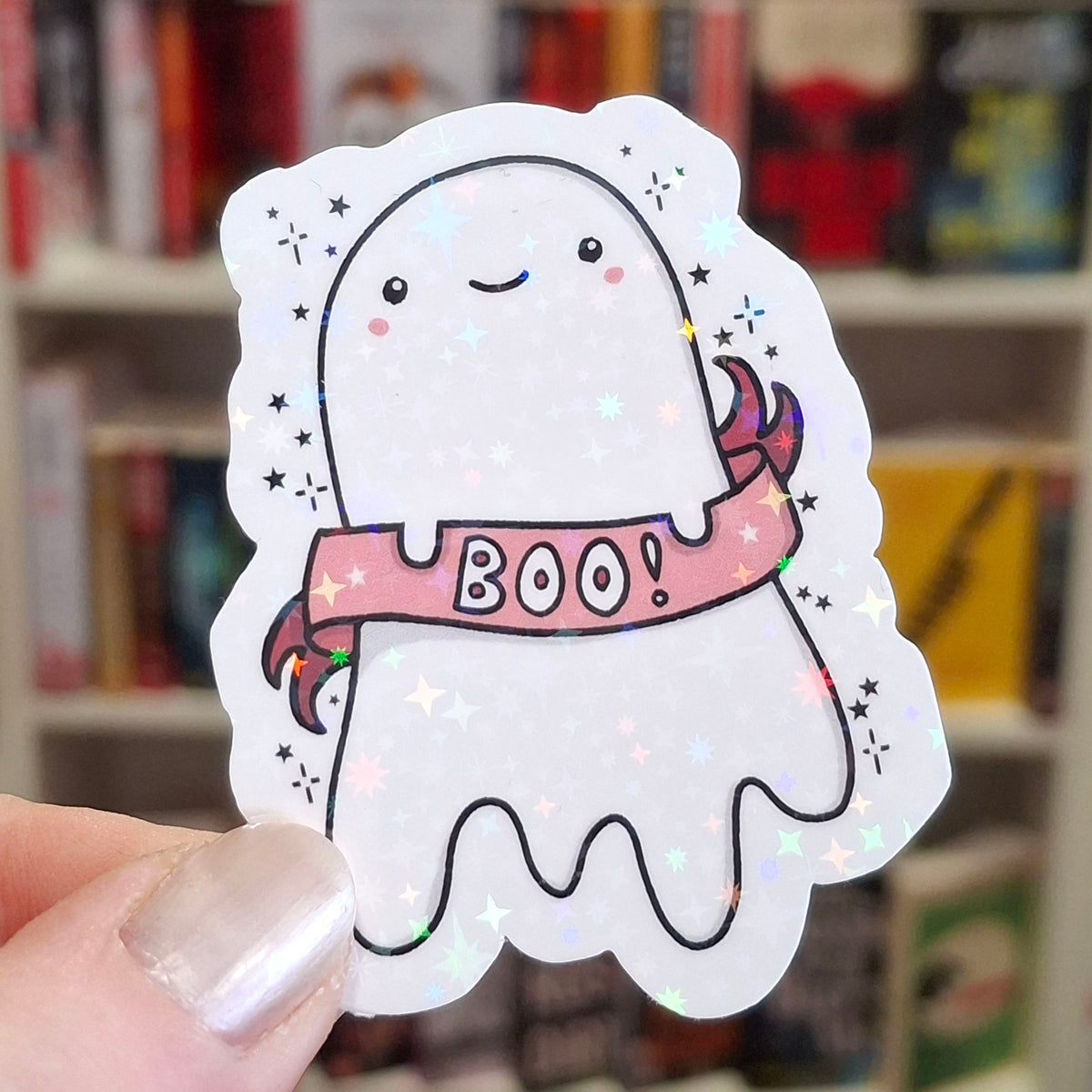 A close-up of a hand holding a die-cut holographic sticker featuring a cute white cartoon ghost holding a pink 'BOO!' banner, with a blurred bookshelf in the background.