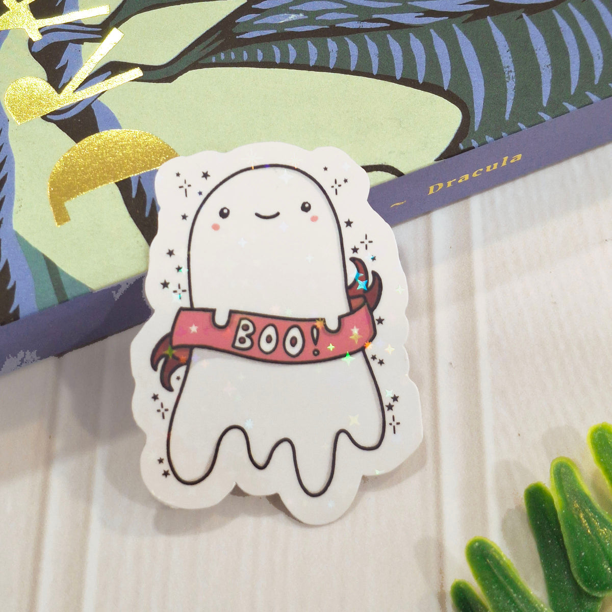 A cute holographic ghost sticker with a pink 'BOO!' banner, placed on a light wood surface next to a decorative copy of the book Dracula.