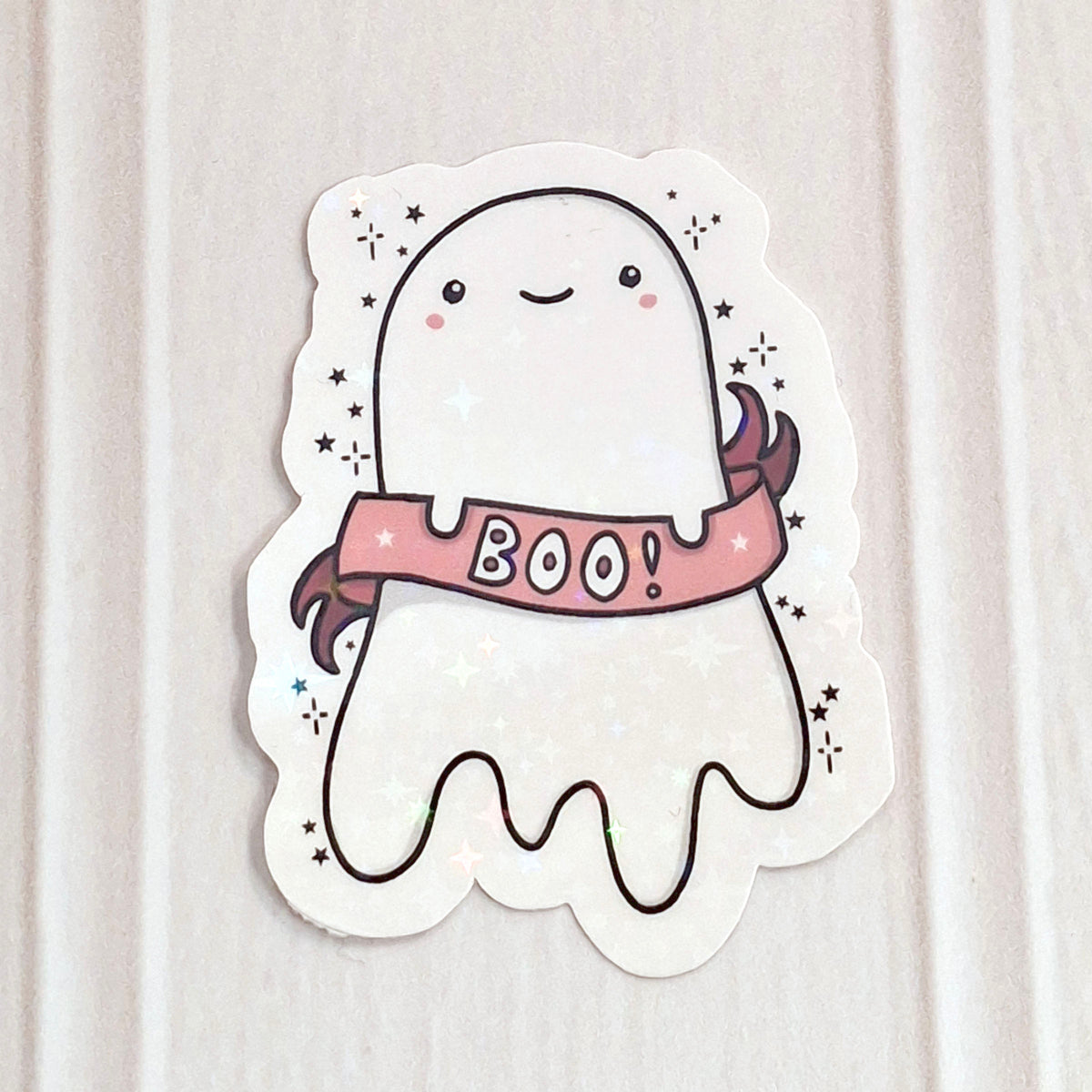 A cute holographic ghost shaped sticker with a pink 'BOO!' banner, shown on a light wood background.