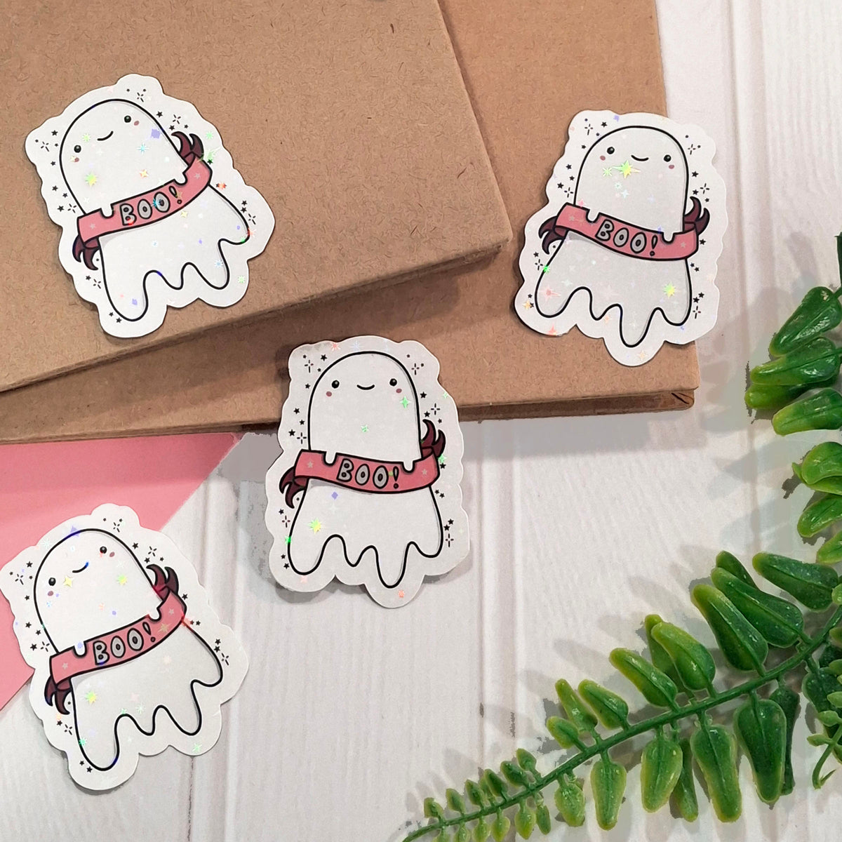 Four cute holographic ghost stickers with pink 'BOO!' banners are arranged on brown kraft notebooks and a white wood surface with a green plant leaf in the corner.