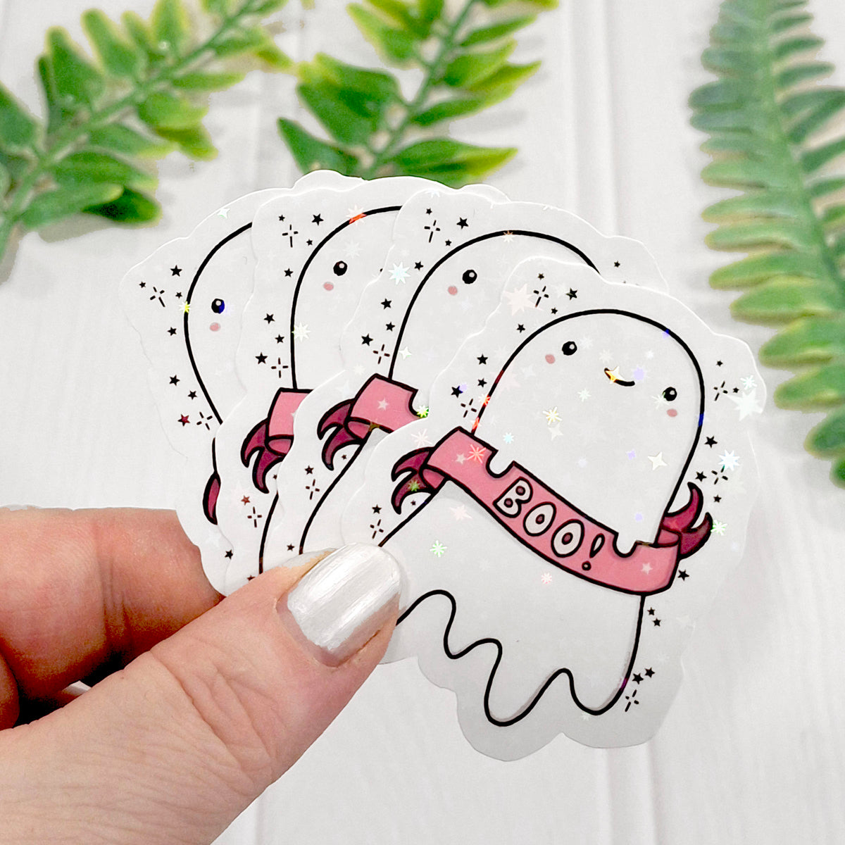 A hand holds a stack of four die-cut holographic stickers featuring cute cartoon ghosts with pink banners that say 'BOO!'. The stickers have black outlines, small black stars, and subtle holographic glitter.
