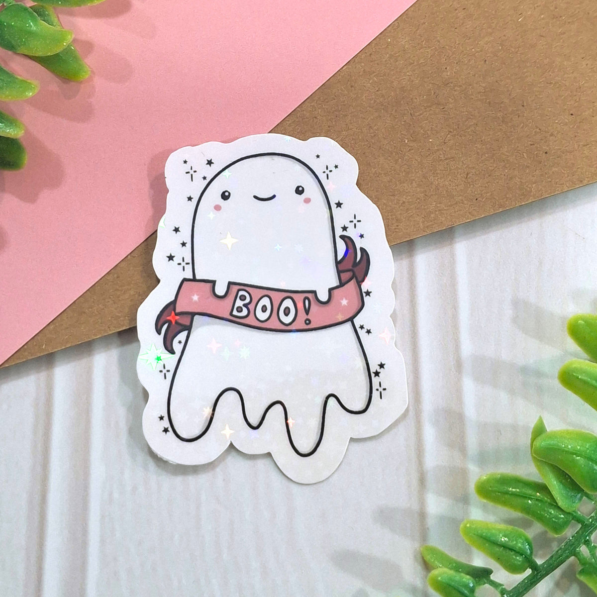 A cute holographic ghost sticker with a pink 'BOO!' banner, placed on a white wood surface with a pink paper background, a kraft brown envelope, and green plant leaves.