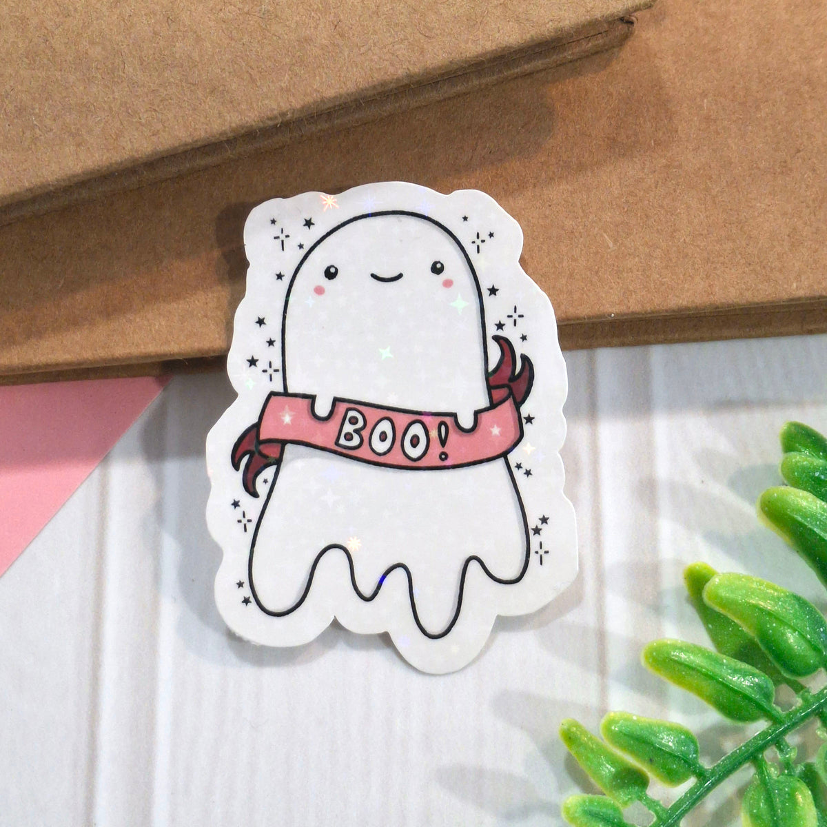 A cute holographic ghost-shaped sticker with a pink 'BOO!' banner, placed on a white surface with a brown kraft notebook and green leaves in the background.