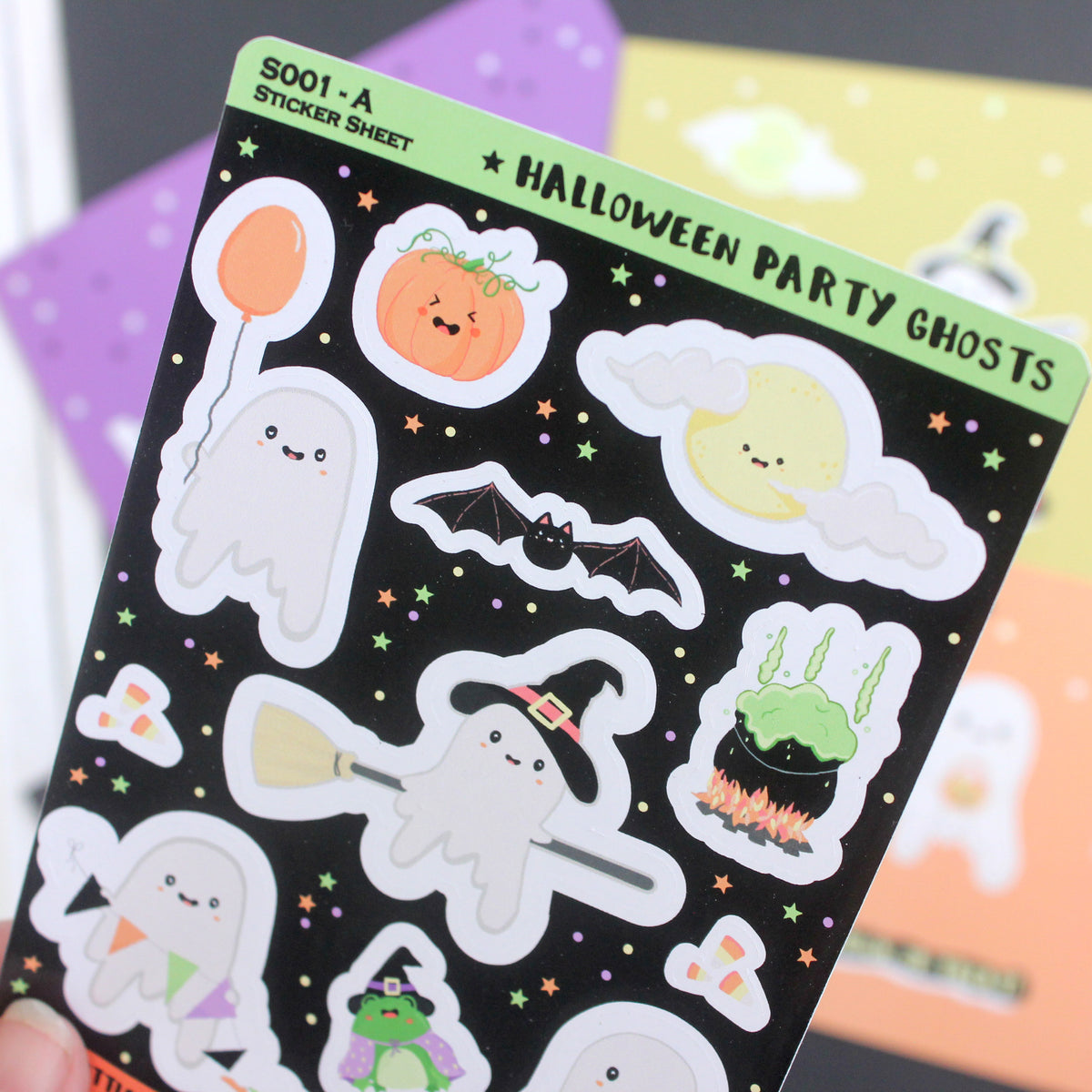 A close-up of a Halloween sticker sheet featuring cute ghost characters, a bat, a pumpkin, a crescent moon, and a cauldron of potion, all on a black background with a green header.