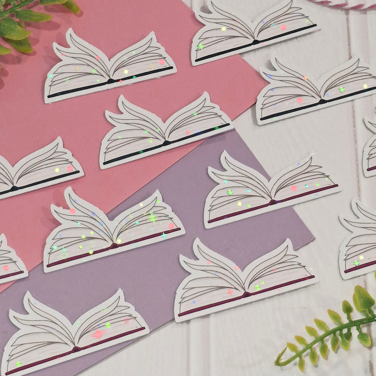 An image showing the detail and sparkles of the holographic open fluttering book pages embellishments, arranged on a pink and lavender background with a green leaf in the corner.
