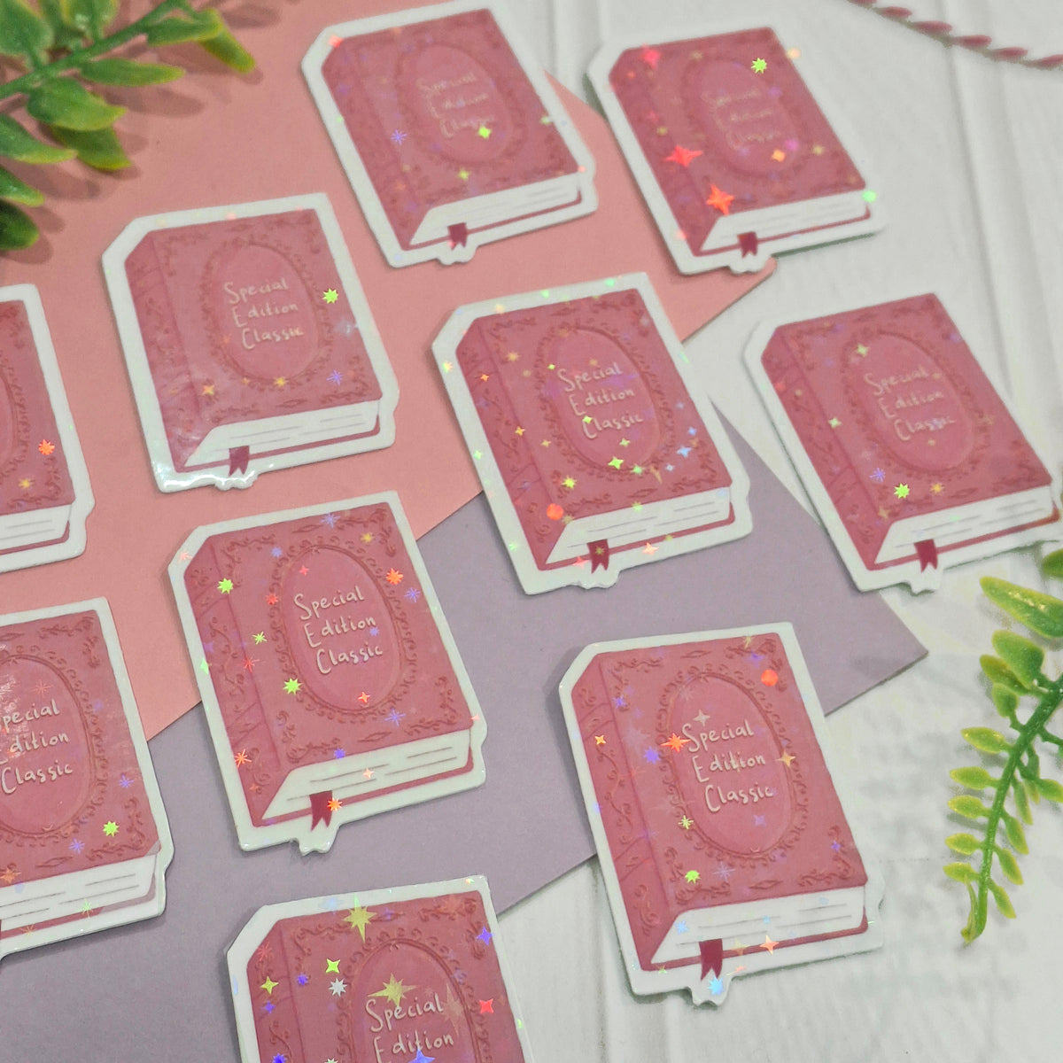 An image showing the detail and sparkles of the holographic pink book cover embellishments, arranged on a pink and lavender background with a green leaf in the corner.