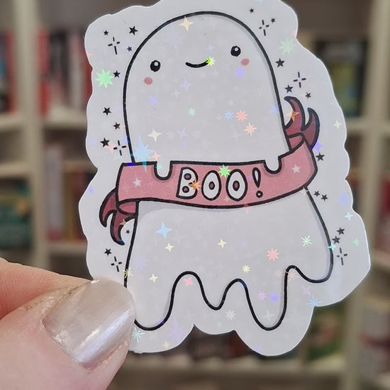 A cute holographic ghost-shaped sticker with a pink 'BOO!' banner, is held in front of blurred book cases. The hand turns the sticker a few different ways and the holographic stars sparkle and shine brightly.