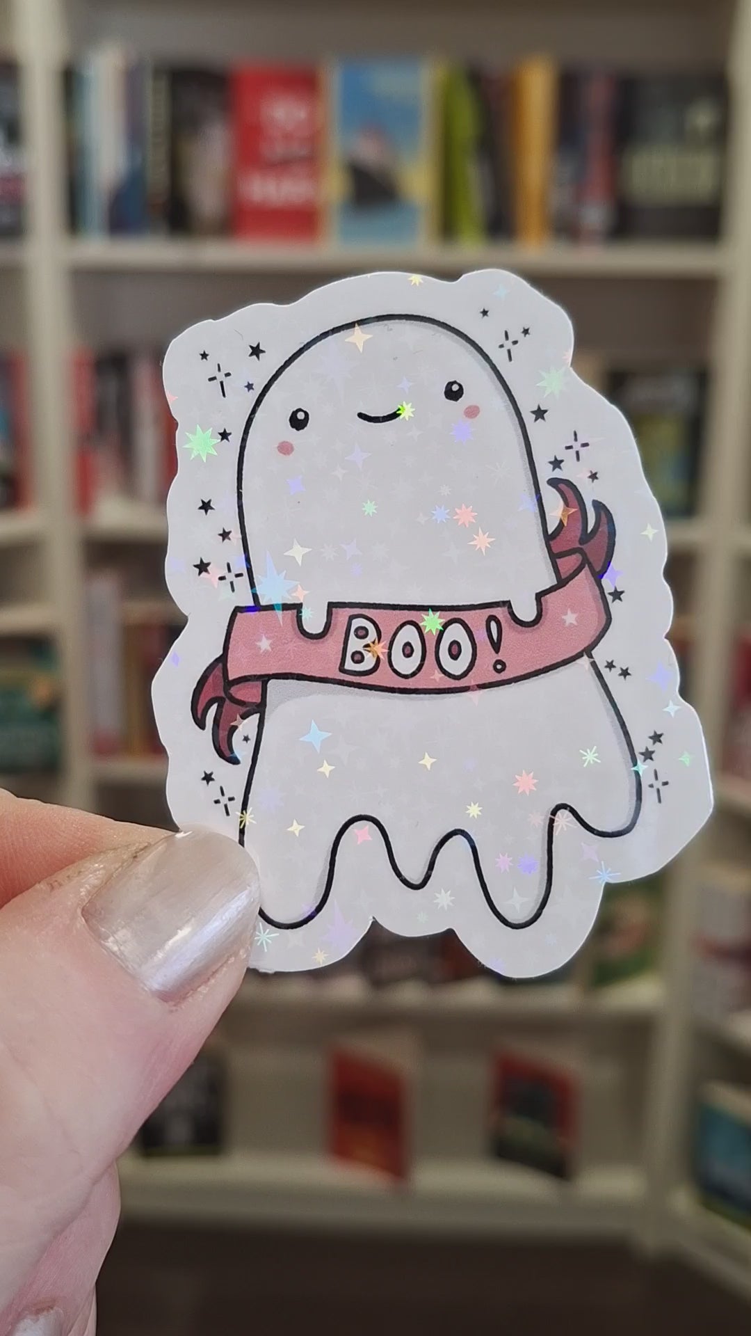 A cute holographic ghost-shaped sticker with a pink 'BOO!' banner, is held in front of blurred book cases. The hand turns the sticker a few different ways and the holographic stars sparkle and shine brightly.