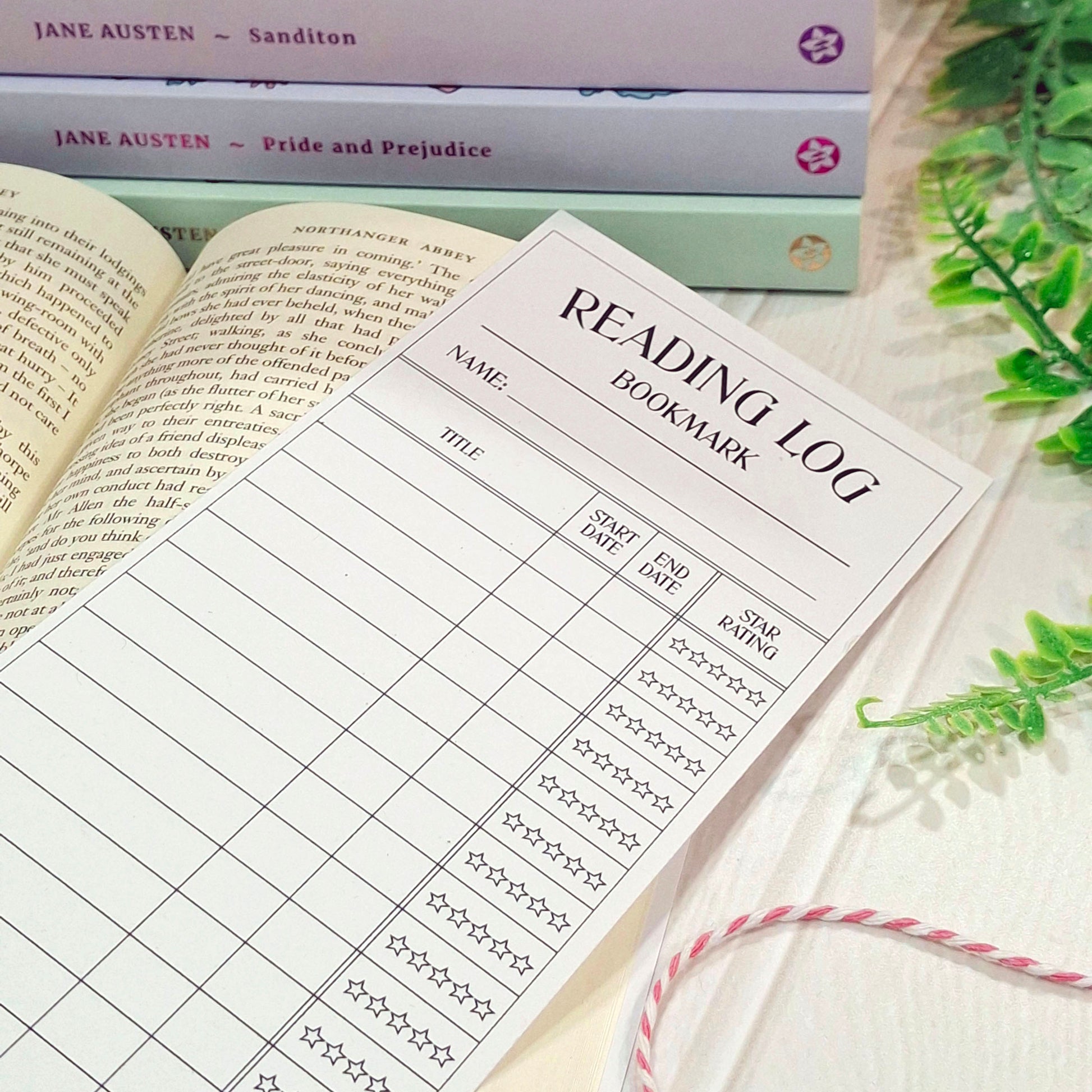 A printed reading log bookmark sheet laid over an open book, with stacked Jane Austen books in the background and green leaves at the side on a light surface.