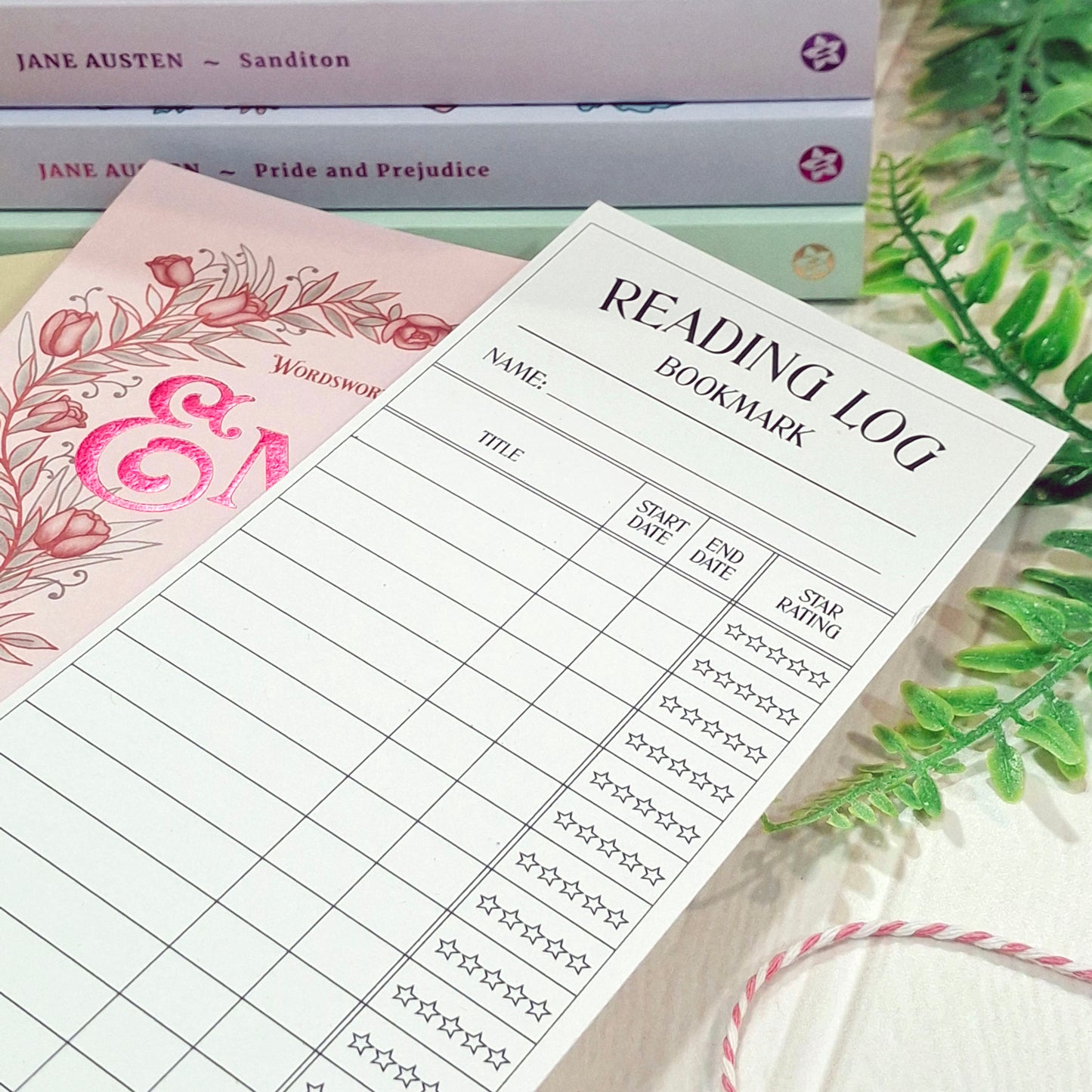 A white reading bookmark log sheet with black printed tables and headings, styled on top of a pink floral book, with books and green leaves in the background.