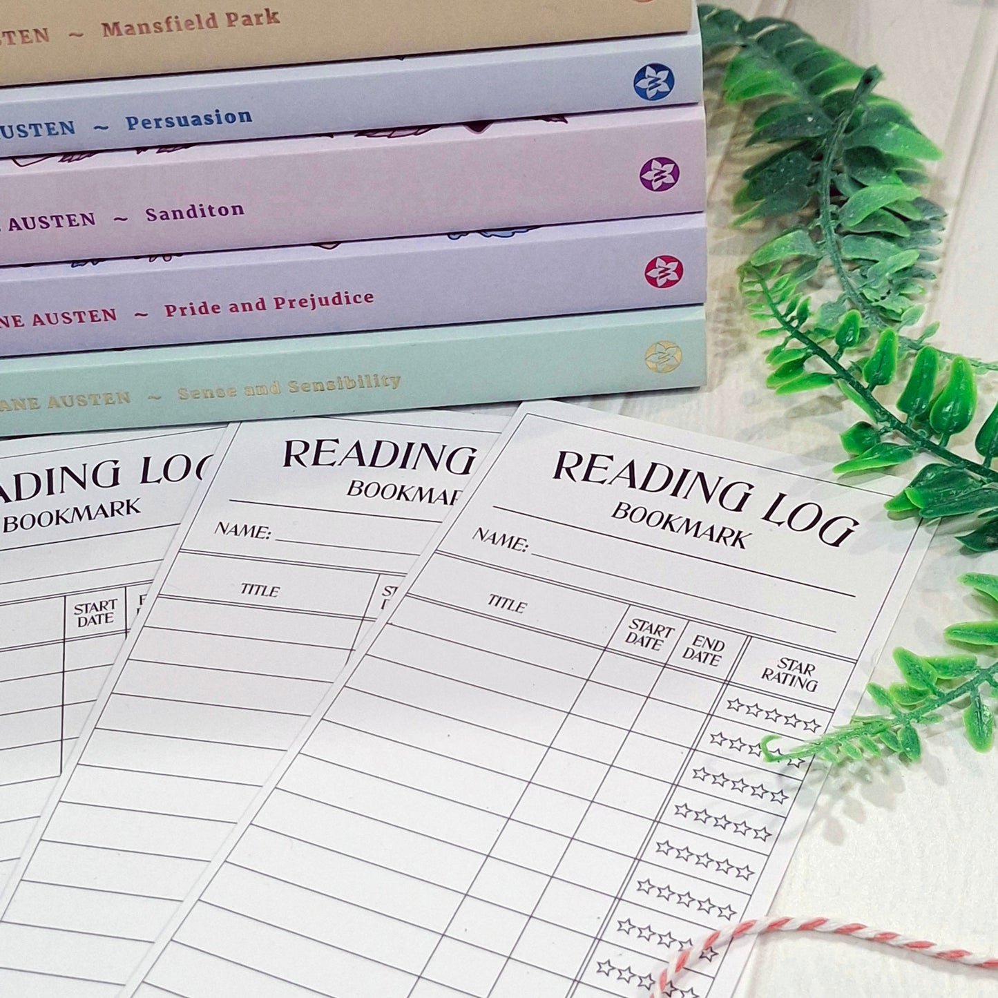 Three printed reading log bookmark sheets laid on a light surface, with a stack of books in the background and green leaves at the side. The sheets are labeled 'READING LOG BOOKMARK' and include fields for name, title, start date, end date, and star rating.