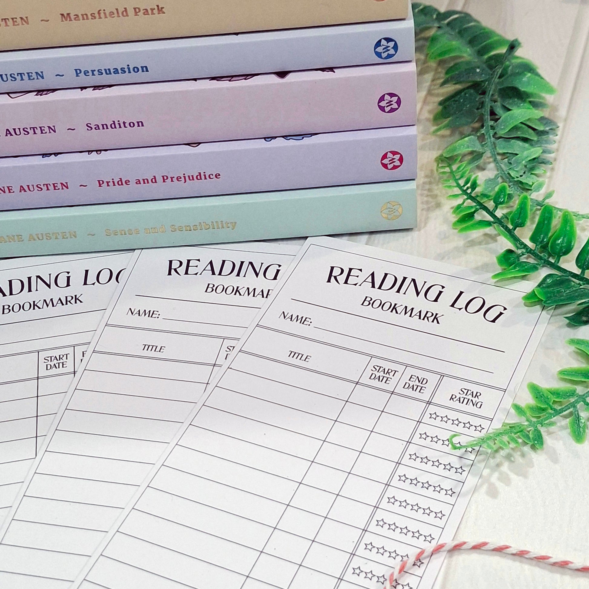 Three printed reading log bookmark sheets laid on a light surface, with a stack of books in the background and green leaves at the side. The sheets are labeled 'READING LOG BOOKMARK' and include fields for name, title, start date, end date, and star rating.