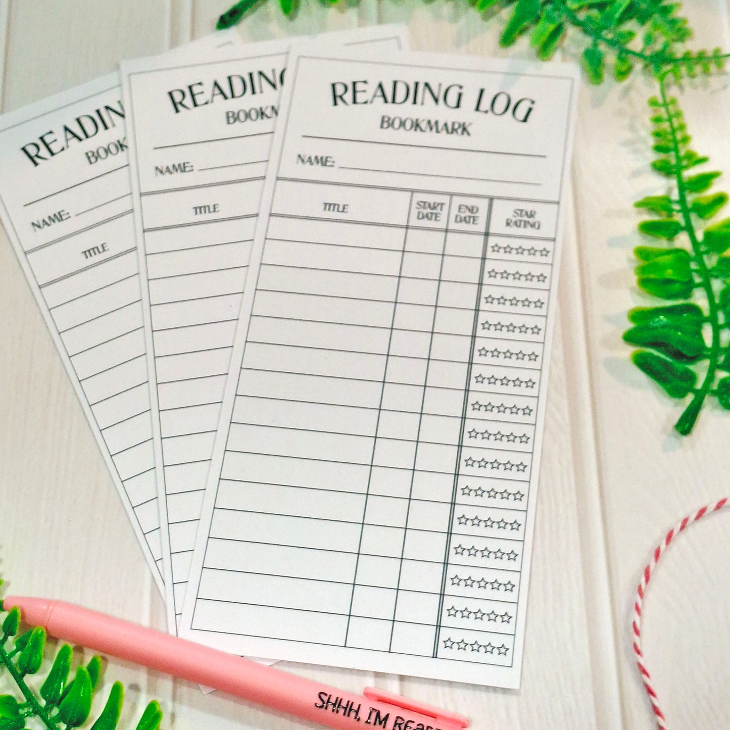 Three printed reading log bookmark sheets arranged on a light wood surface, with a pink pen and green leaf props around them.