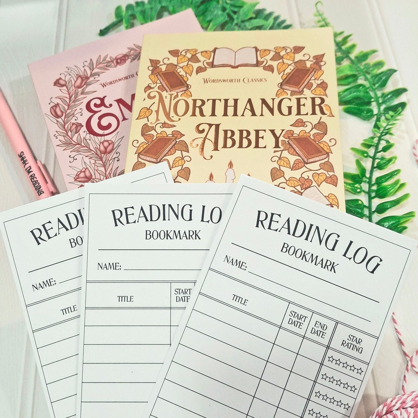 A styled flat-lay of reading-themed stationery and book covers. In the foreground are three white 'Reading Log Bookmark' sheets with black printed lines and text.