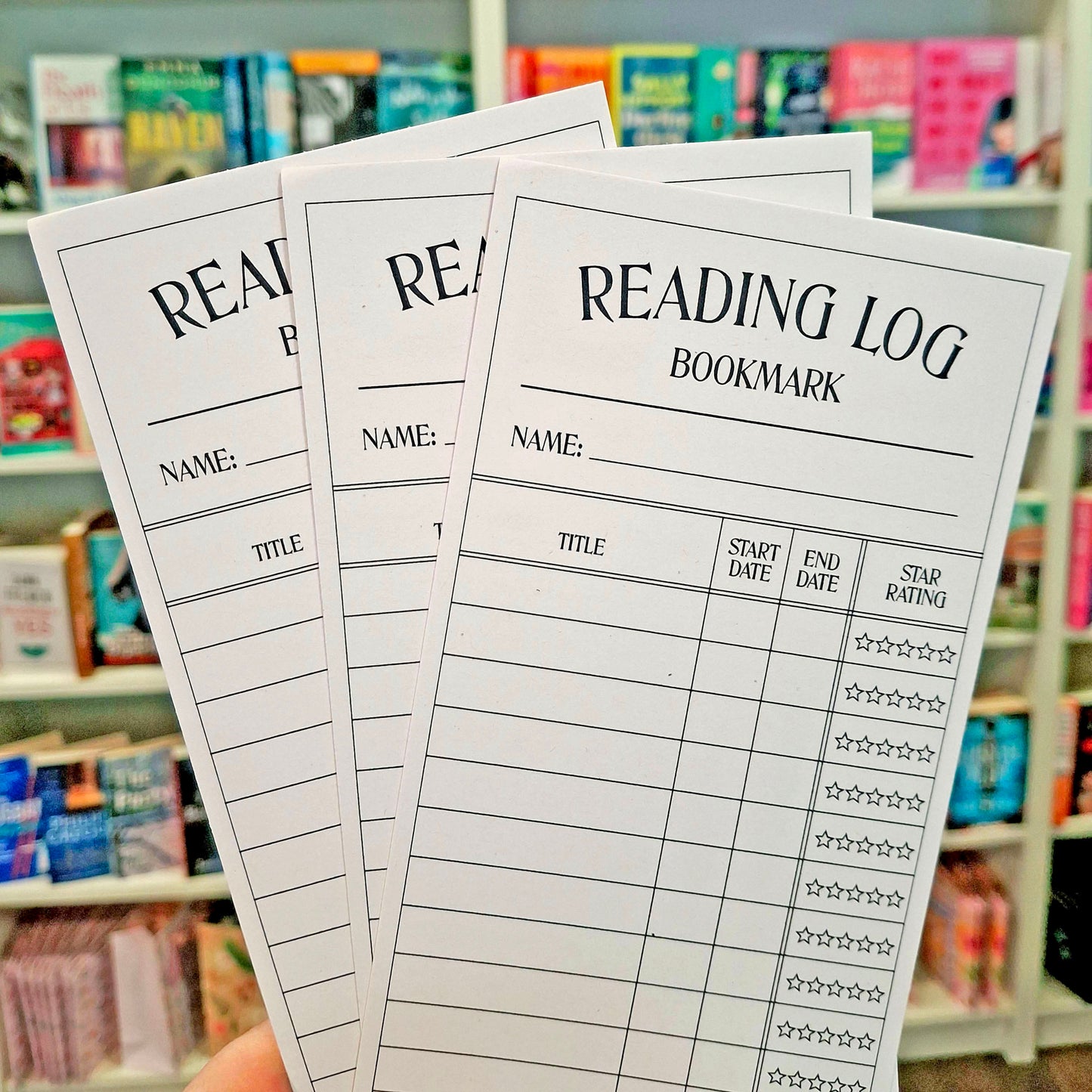 Three printed reading log bookmark sheets are fanned out in front of a blurred bookshelf background. The sheets have black text reading 'READING LOG BOOKMARK' and fields for title, start date, end date, and star rating.