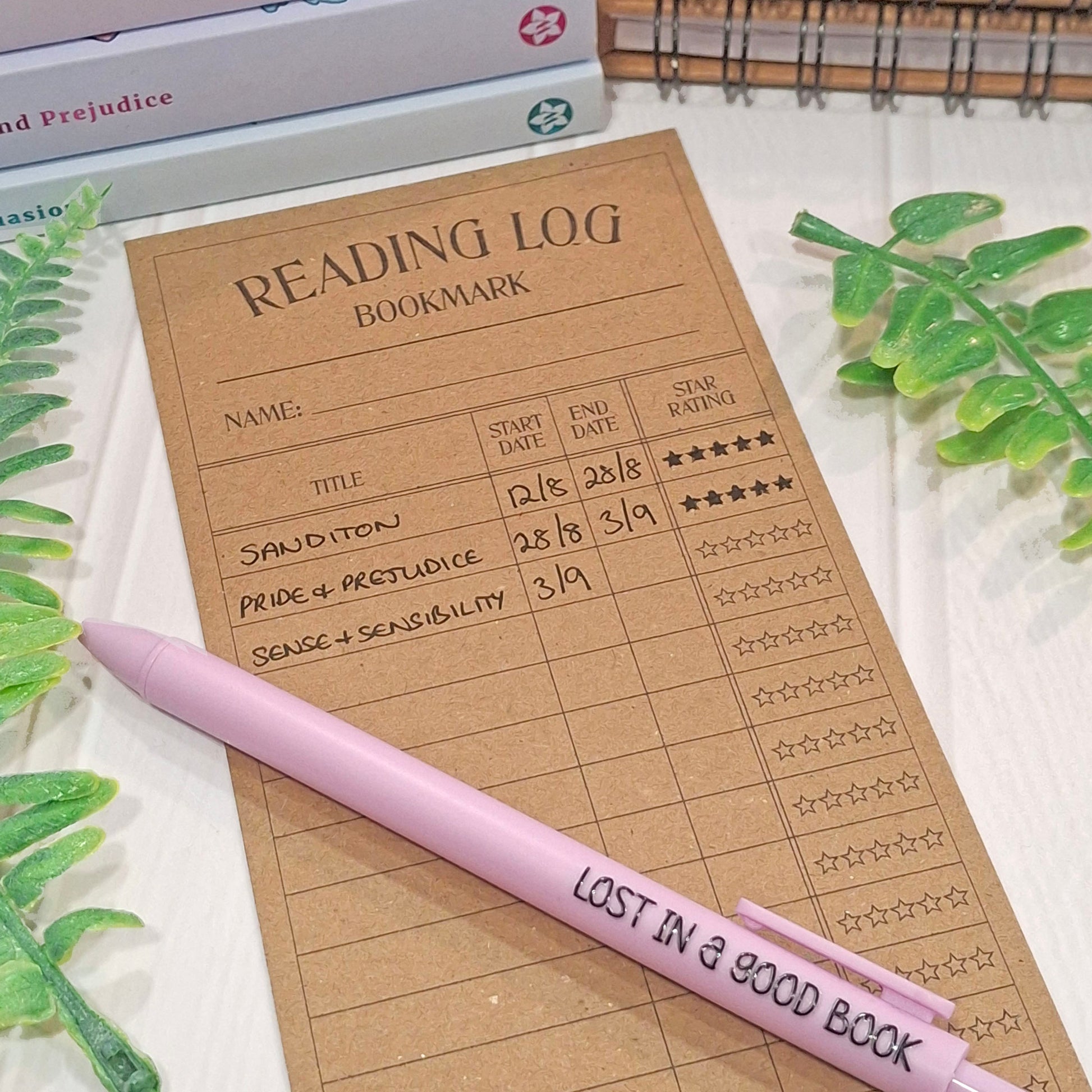 A kraft brown reading log bookmark with a light purple pen placed on top, styled on a white surface with green leaves and books in the background.