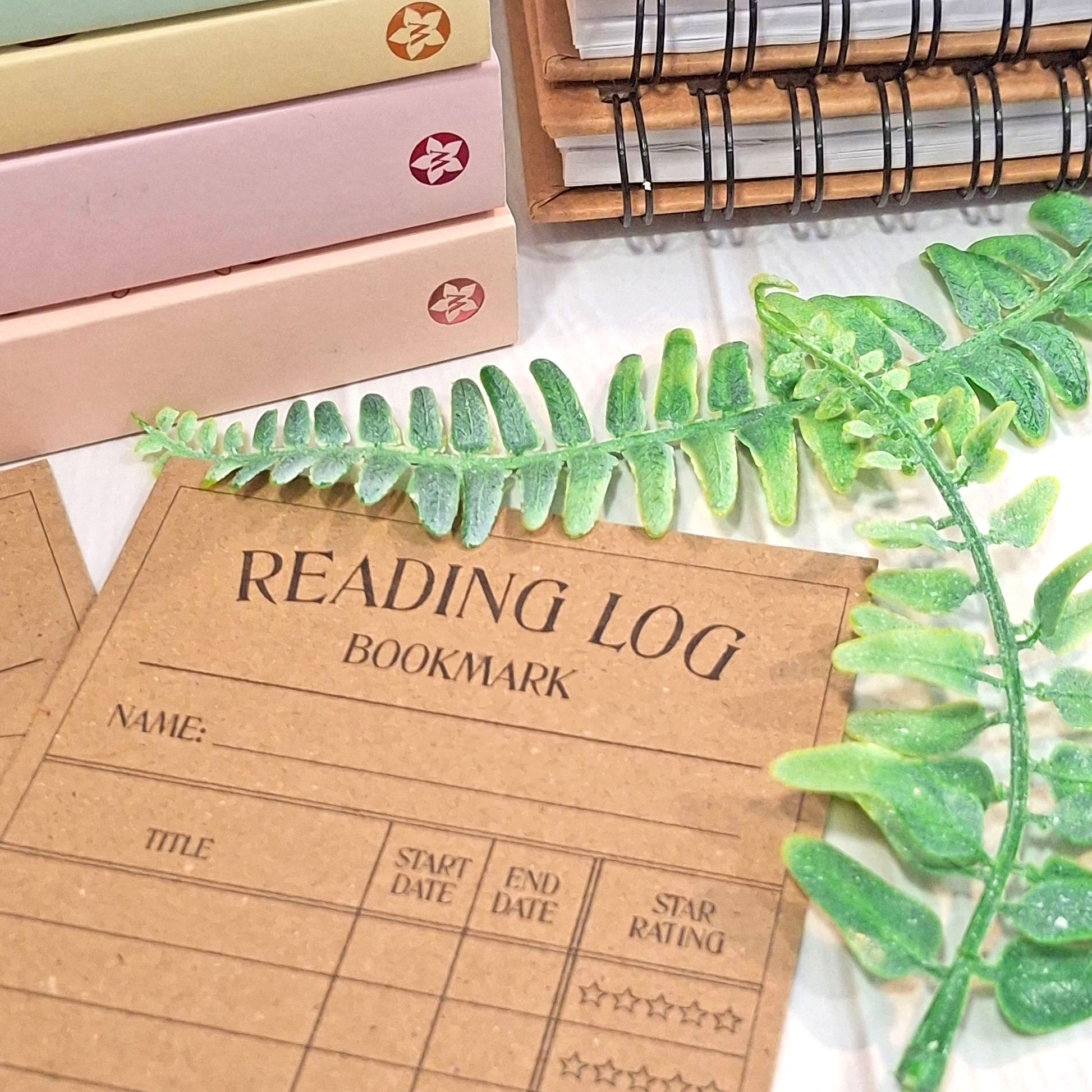 A brown kraft card reading log bookmark with printed fields for name, title, start date, end date, and star rating, styled on a tabletop with a green fern sprig, stacked pastel books, and a spiral notebook.