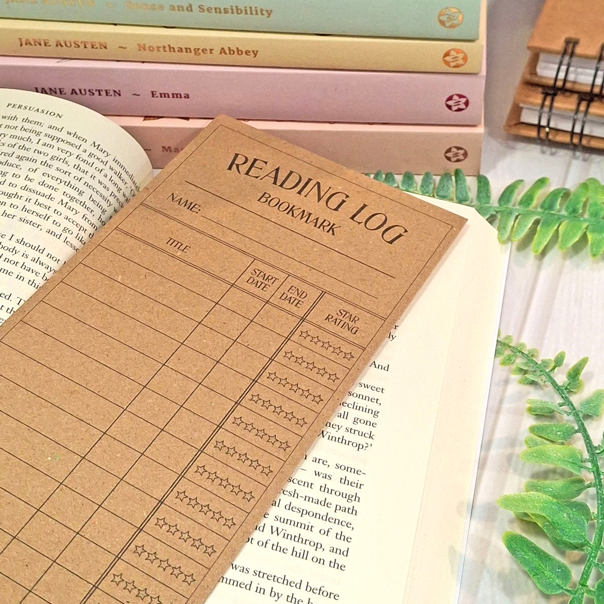 A brown kraft card reading log bookmark styled on an open book, with other books in the background and green leaves on a white surface.