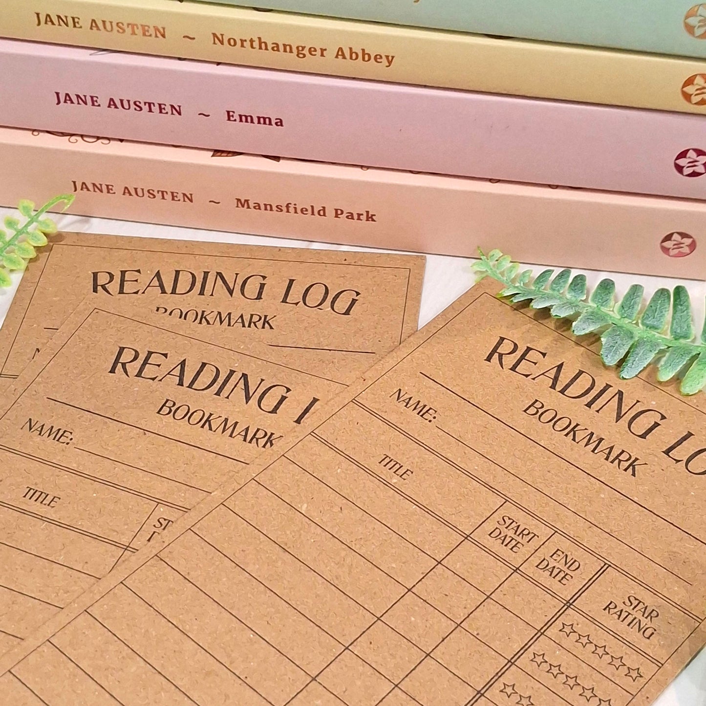Brown kraft card reading log bookmarks with printed fields for title, name, start and end dates, and a star rating, arranged on top of stacked Jane Austen books with green leaves as styling props.