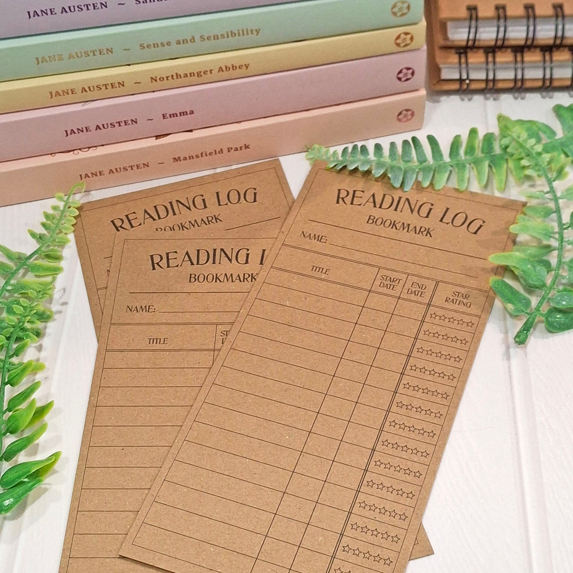 Three brown kraft card reading log bookmark sheets with printed fields and star icons, styled on a white surface with books, green leaves, and a spiral notebook in the background.