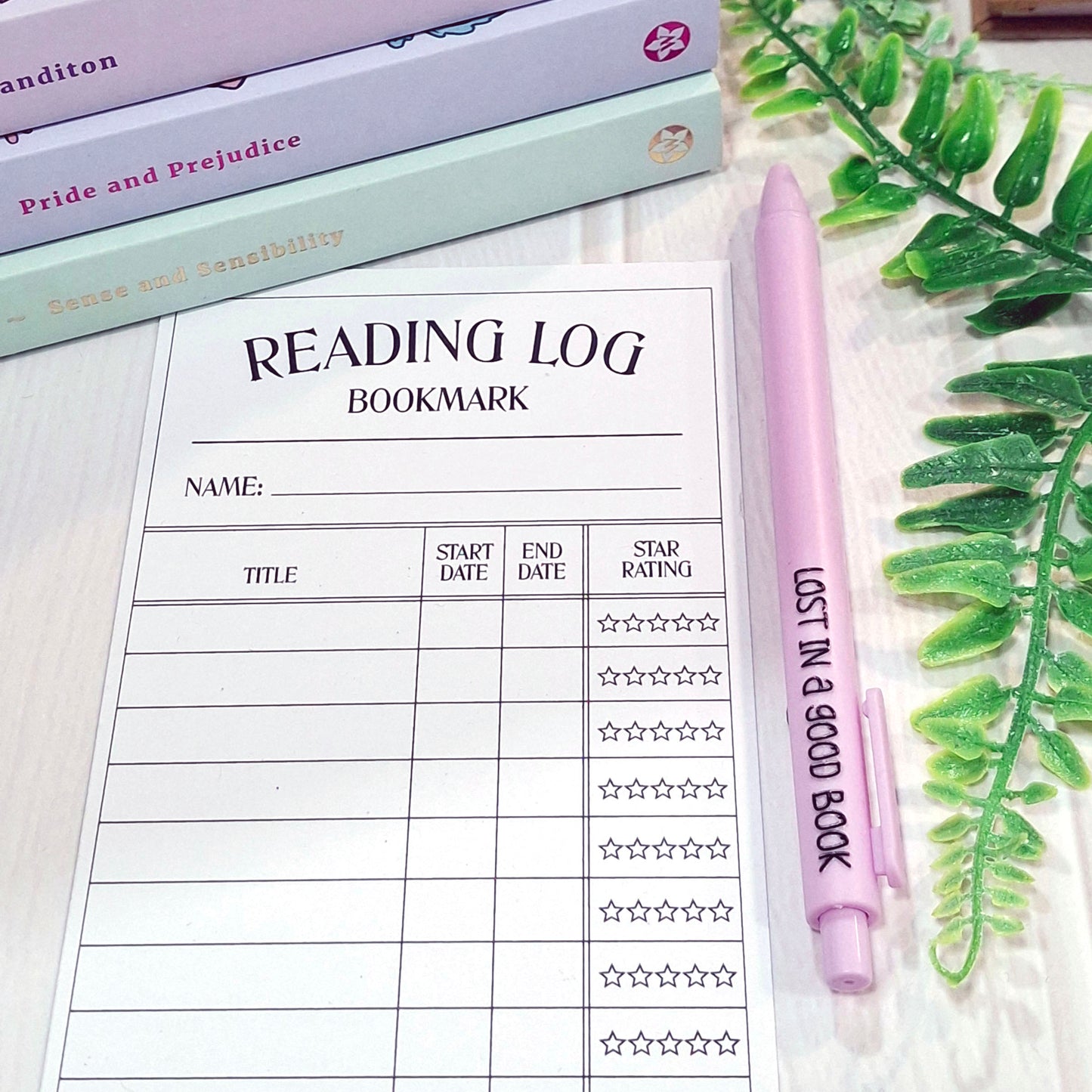 A reading log bookmark sheet with a purple pen labelled 'LOST IN A GOOD BOOK' placed on a light wood surface, styled with stacked books and green leaves.