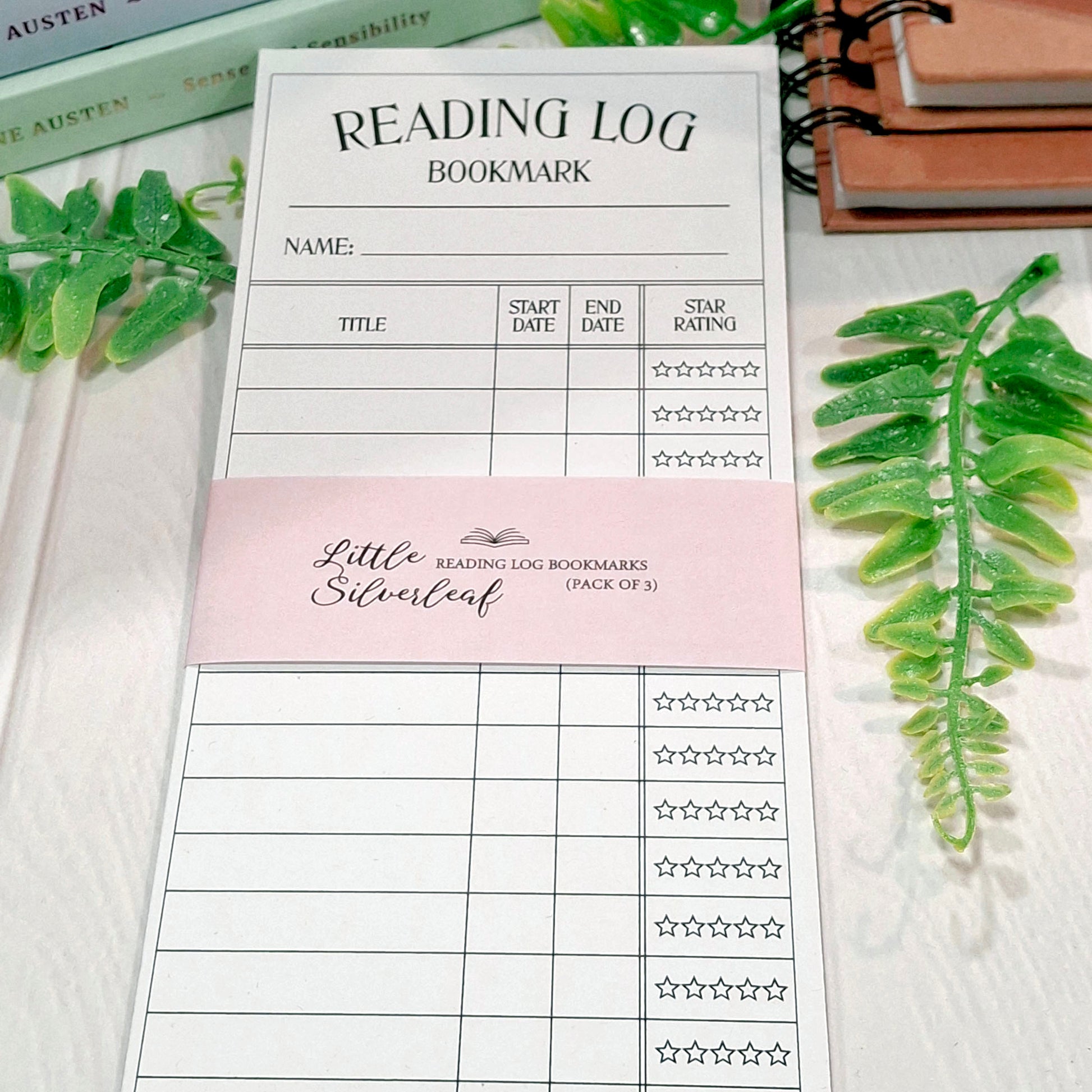 A reading log bookmark set of  3 displayed on a white wooden surface, styled with green leaves and stationery props. The set includes 3 large white bookmarks with black text and is labelled 'Little Silverleaf Reading Log Bookmarks (Pack of 3)'.