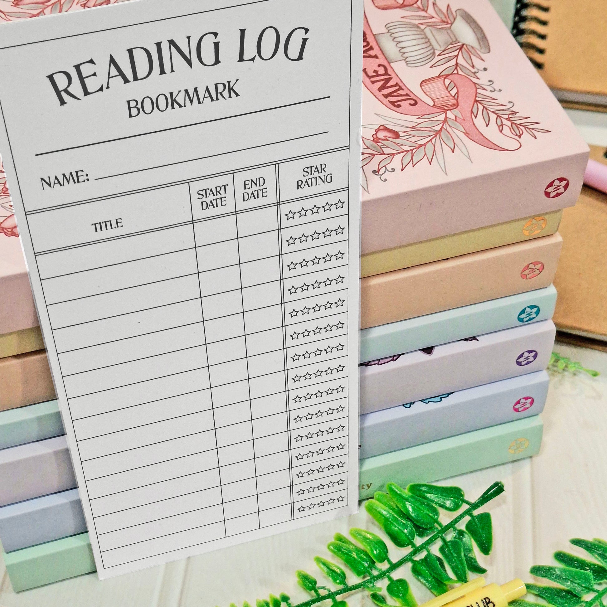 A white reading log bookmark sheet with printed columns for title, start date, end date, and star rating, placed in front of a stack of pastel-coloured books with decorative illustrations.