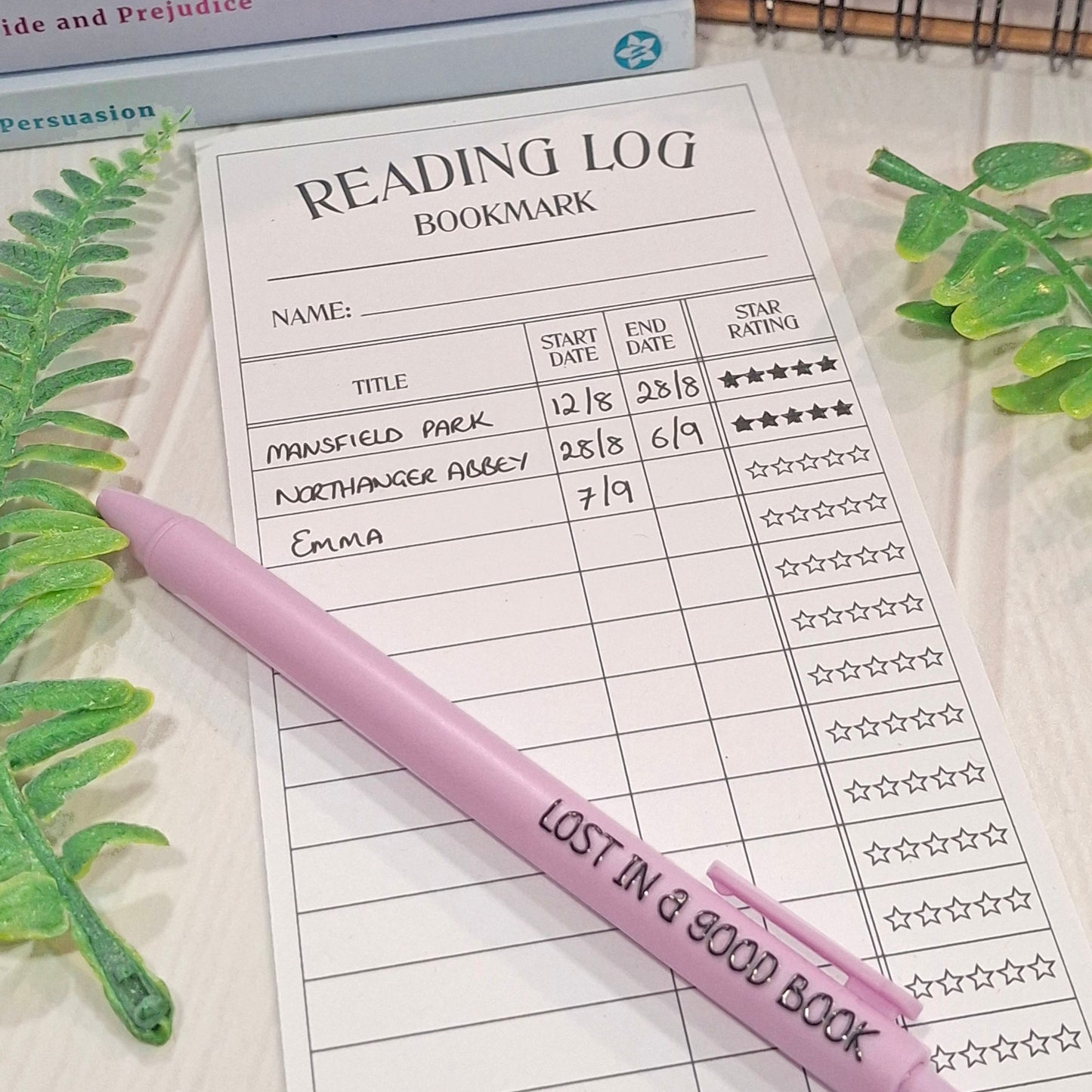 A purple pen with the words 'LOST IN A GOOD BOOK' printed on it, resting on a printed reading log bookmark sheet. The scene is styled with green leaves on a light wood surface.