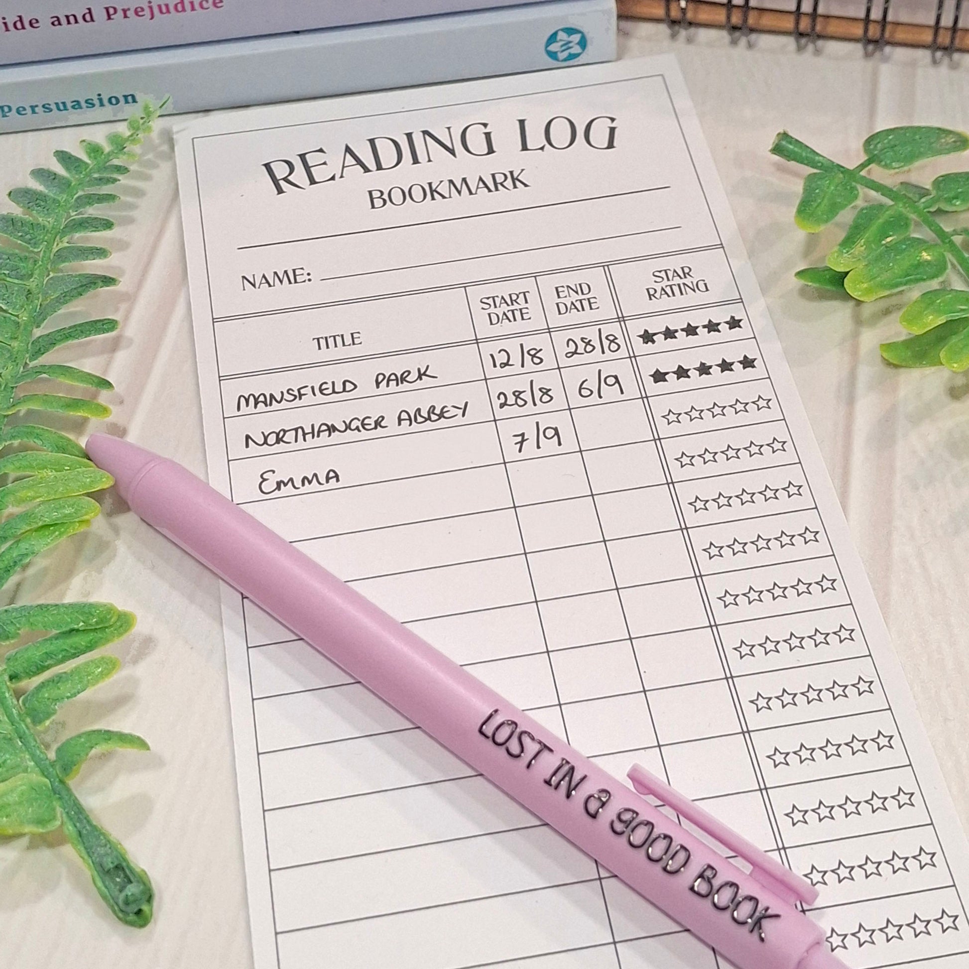 A purple pen with the words 'LOST IN A GOOD BOOK' printed on it, resting on a printed reading log bookmark sheet. The scene is styled with green leaves on a light wood surface.
