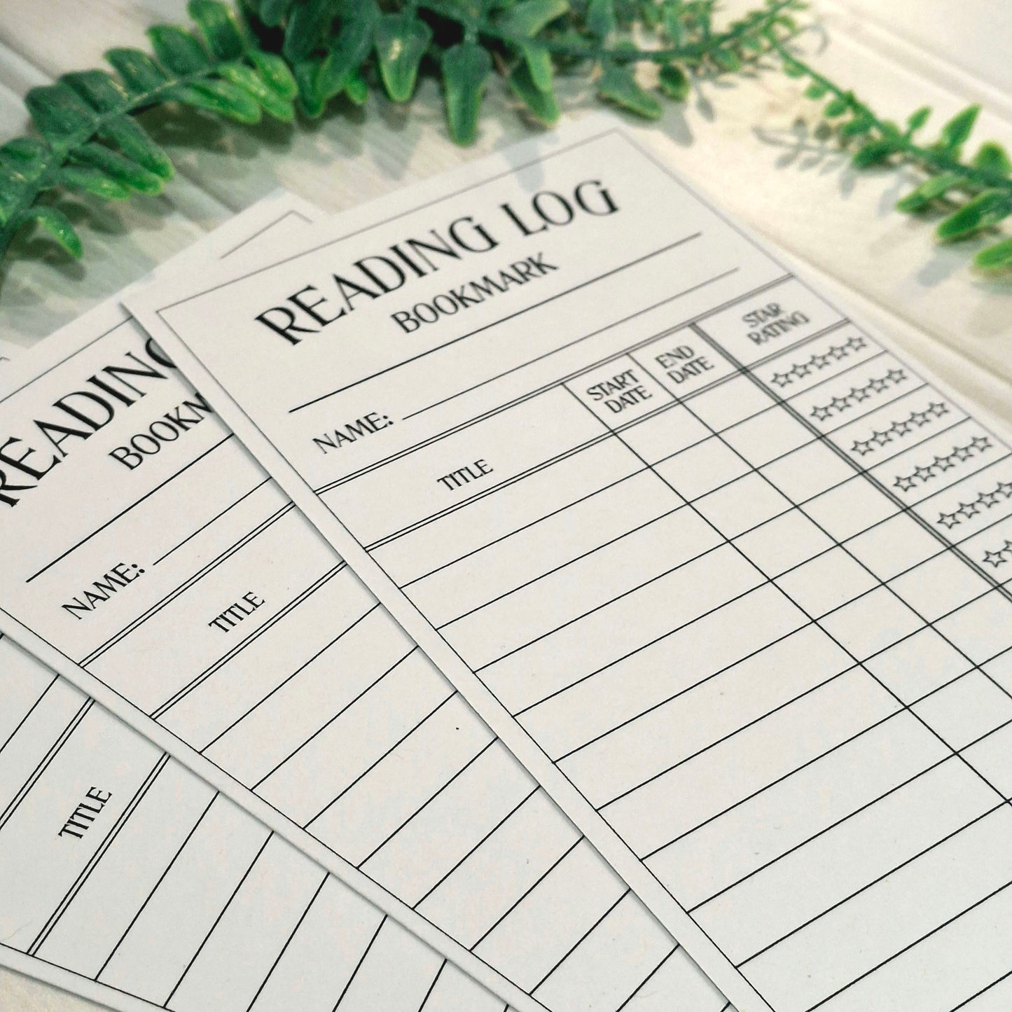 White reading log bookmark sheets with black printed lines and text, arranged on a light surface with green foliage in the background.