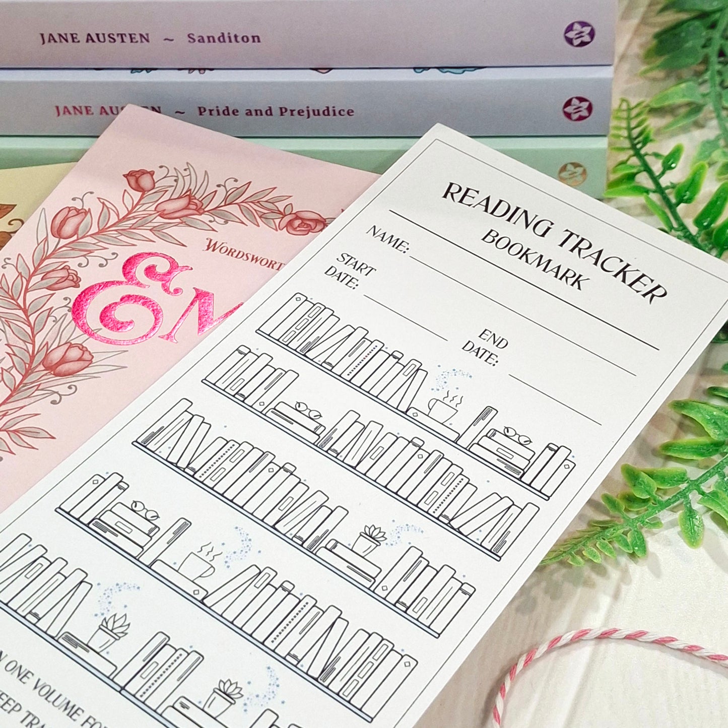 A reading tracker bookmark sheet with black line drawings of bookshelves and fill-in lines, placed over a pink floral book and stacked books in the background.