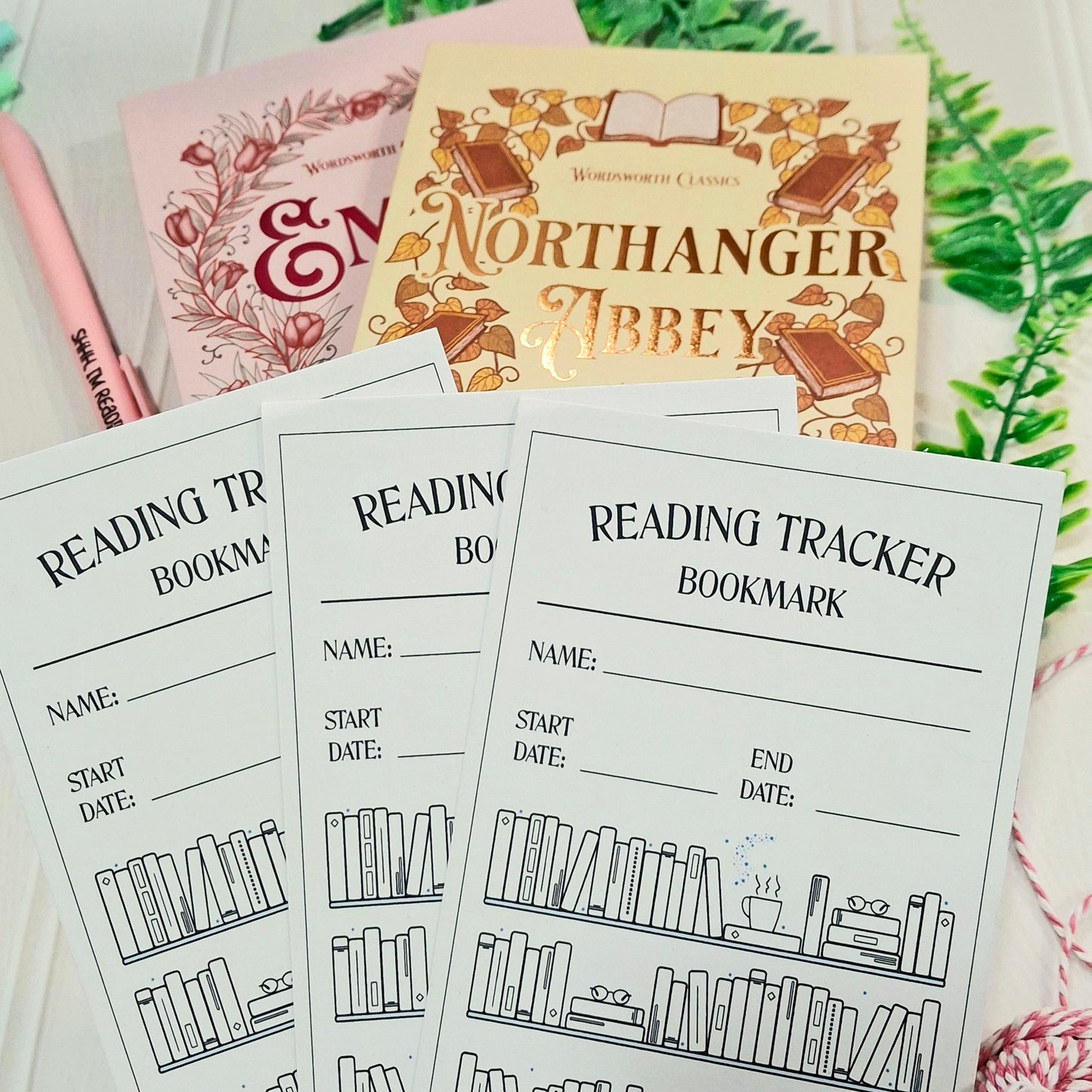 Three printable reading tracker bookmark sheets are arranged on a white surface with a pink book, a gold book, and green leaves in the background.