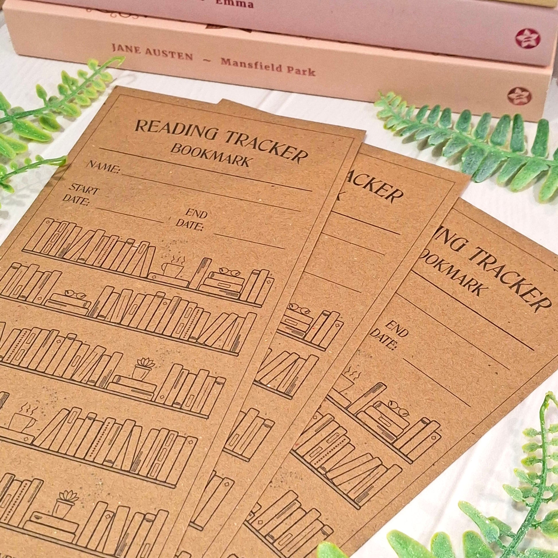 Three brown kraft card reading tracker bookmarks with black printed text and bookshelf illustrations, arranged on a white surface with decorative green leaves and a pink book in the background.