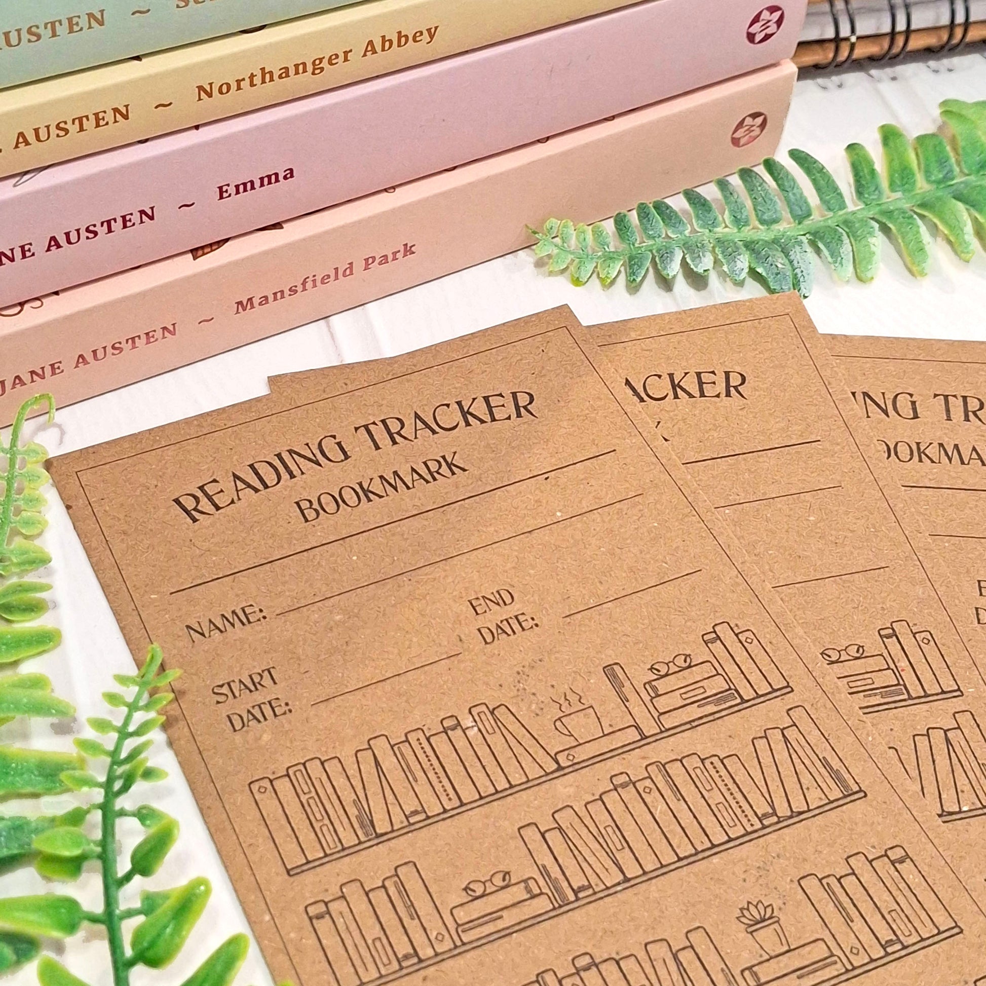 Brown kraft card reading tracker bookmarks with black printed text and bookshelf illustrations, arranged on a white surface with decorative books and green leaves in the background.