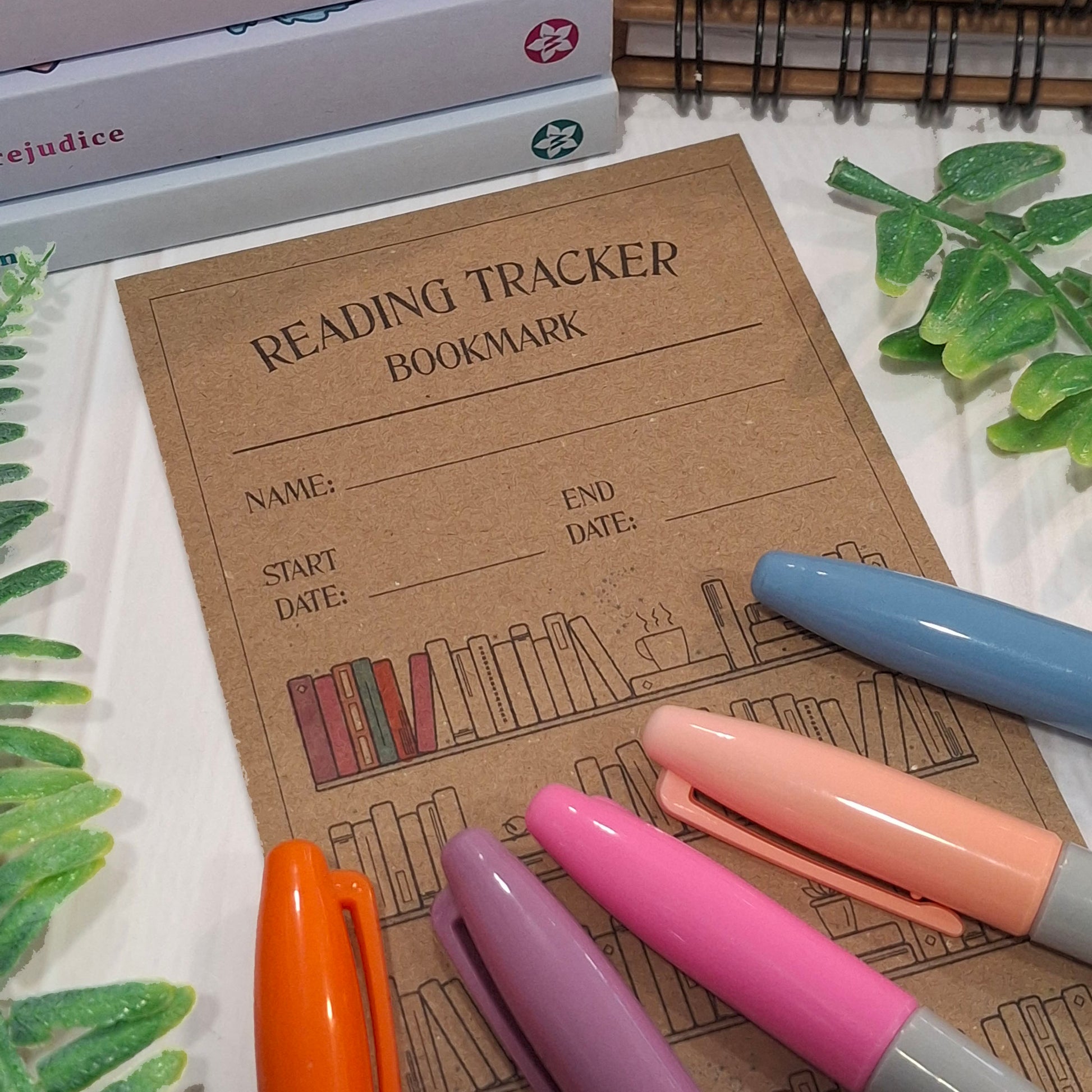 A partly coloured in brown kraft paper reading tracker bookmark with a book illustration and fill-in lines, styled on a white surface with colorful pens and decorative leaves.