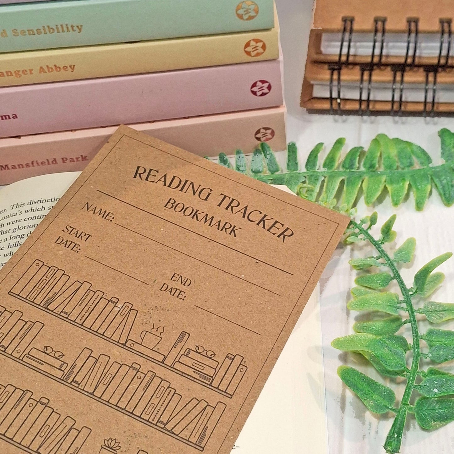 A brown kraft card reading tracker bookmark with black printed text and book illustrations, styled on a tabletop with books, a spiral notebook, and green leaves.
